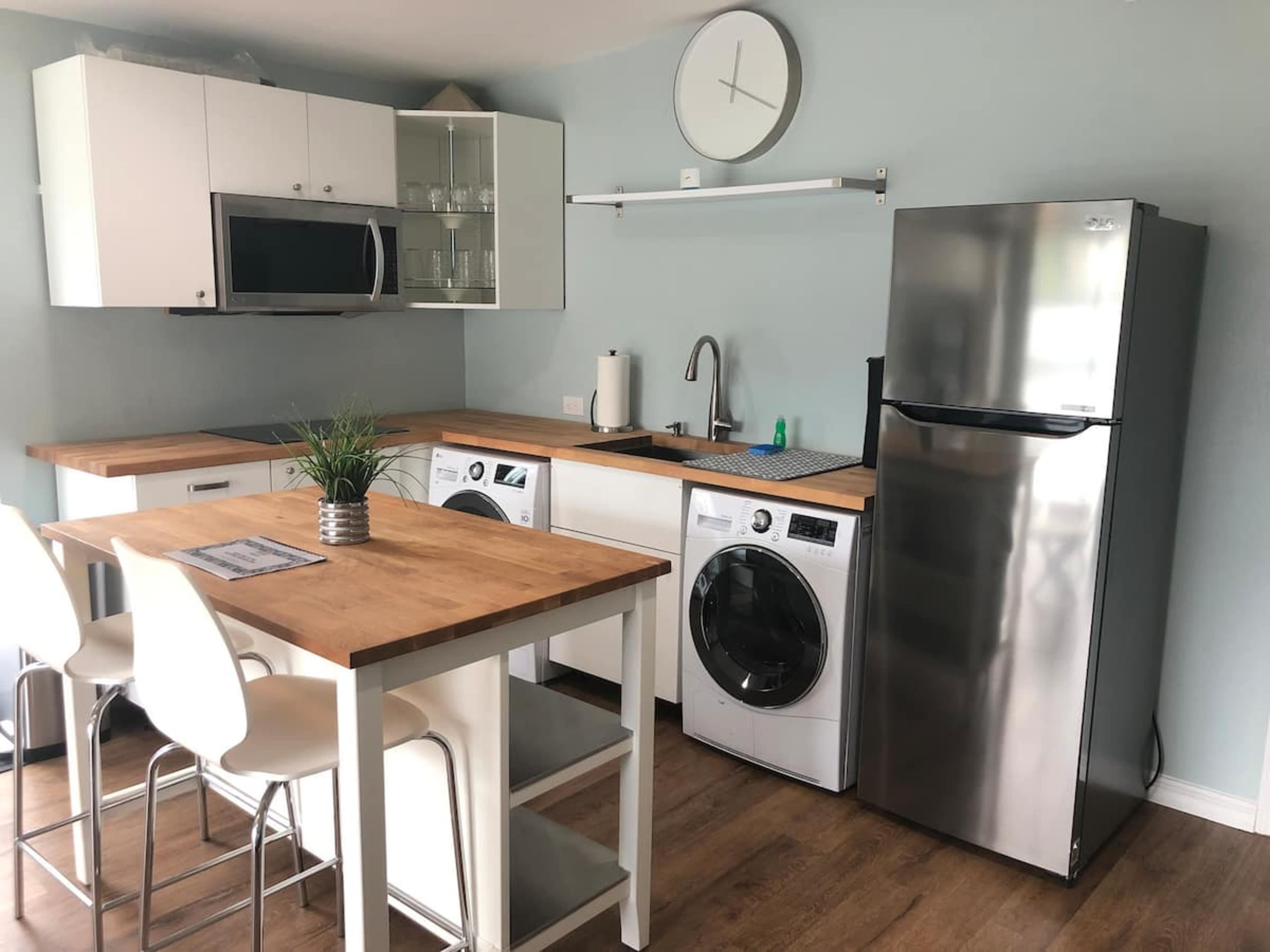 A modern kitchen area equipped with a refrigerator, washer, dryer, microwave, and a wooden dining table with white stools.