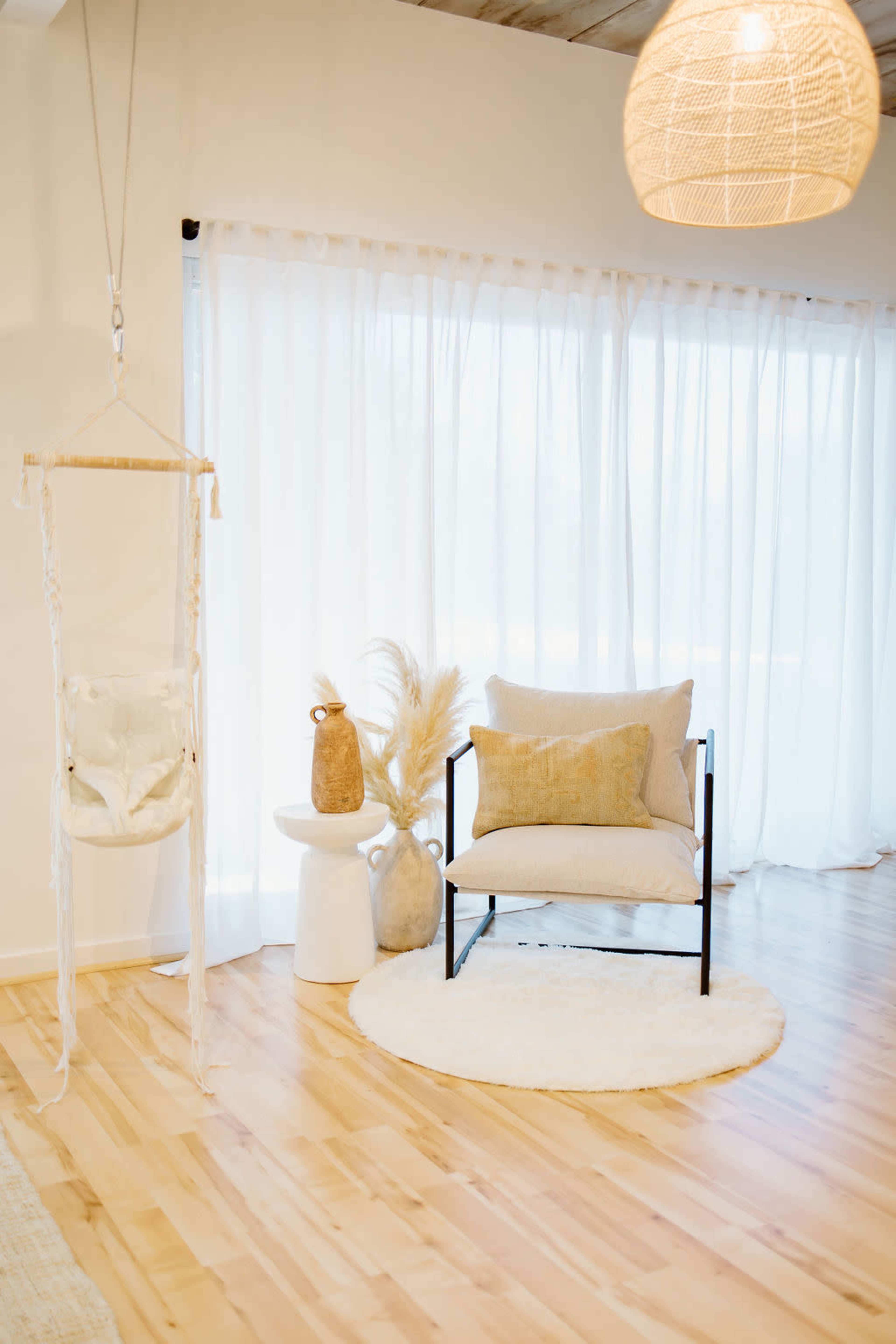 A minimalist room features a black-framed chair with a beige cushion, a small white stool, a decorative vase, and a hanging macrame planter, all set against sheer white curtains.