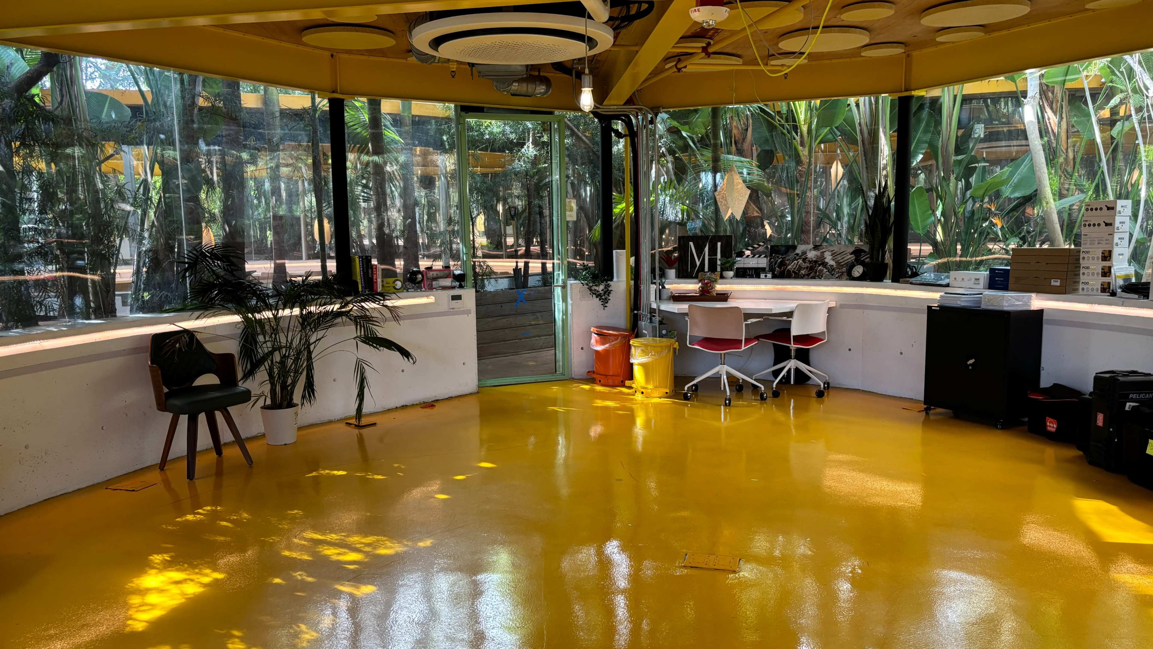 The image shows a bright, open workspace with yellow flooring, large windows, plants, and a table with chairs.