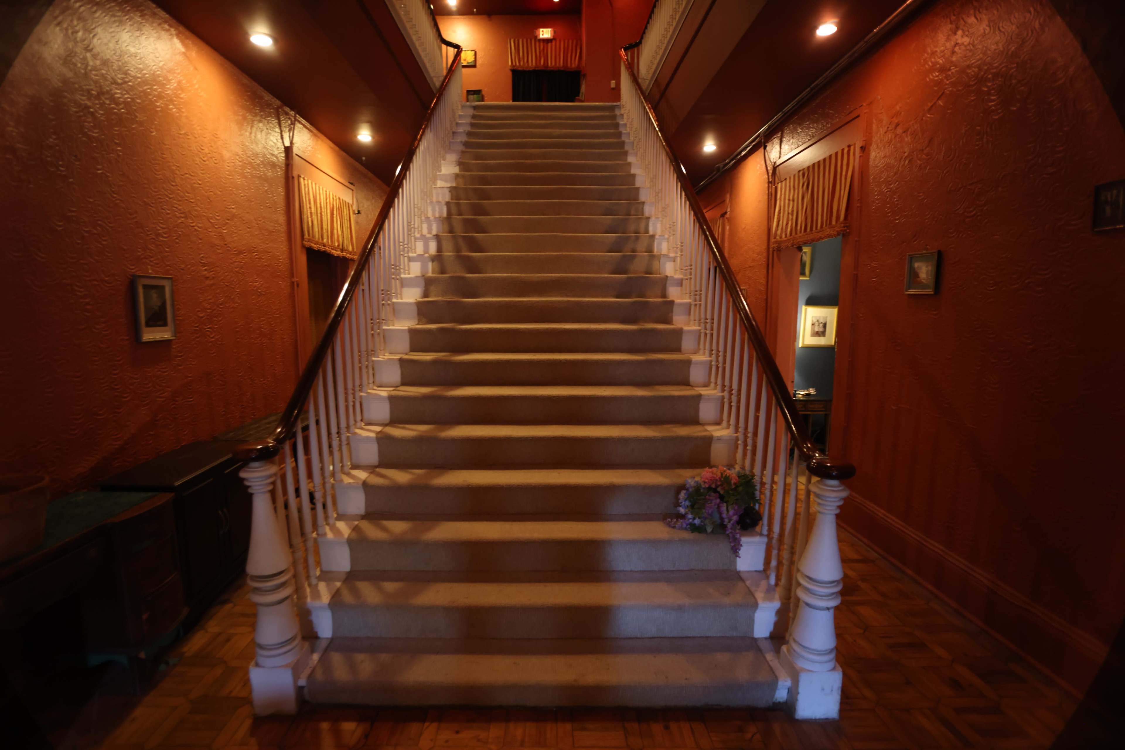 A carpeted staircase with white railings ascends in a hallway lined with textured orange walls and dimly lit by wall sconces.
