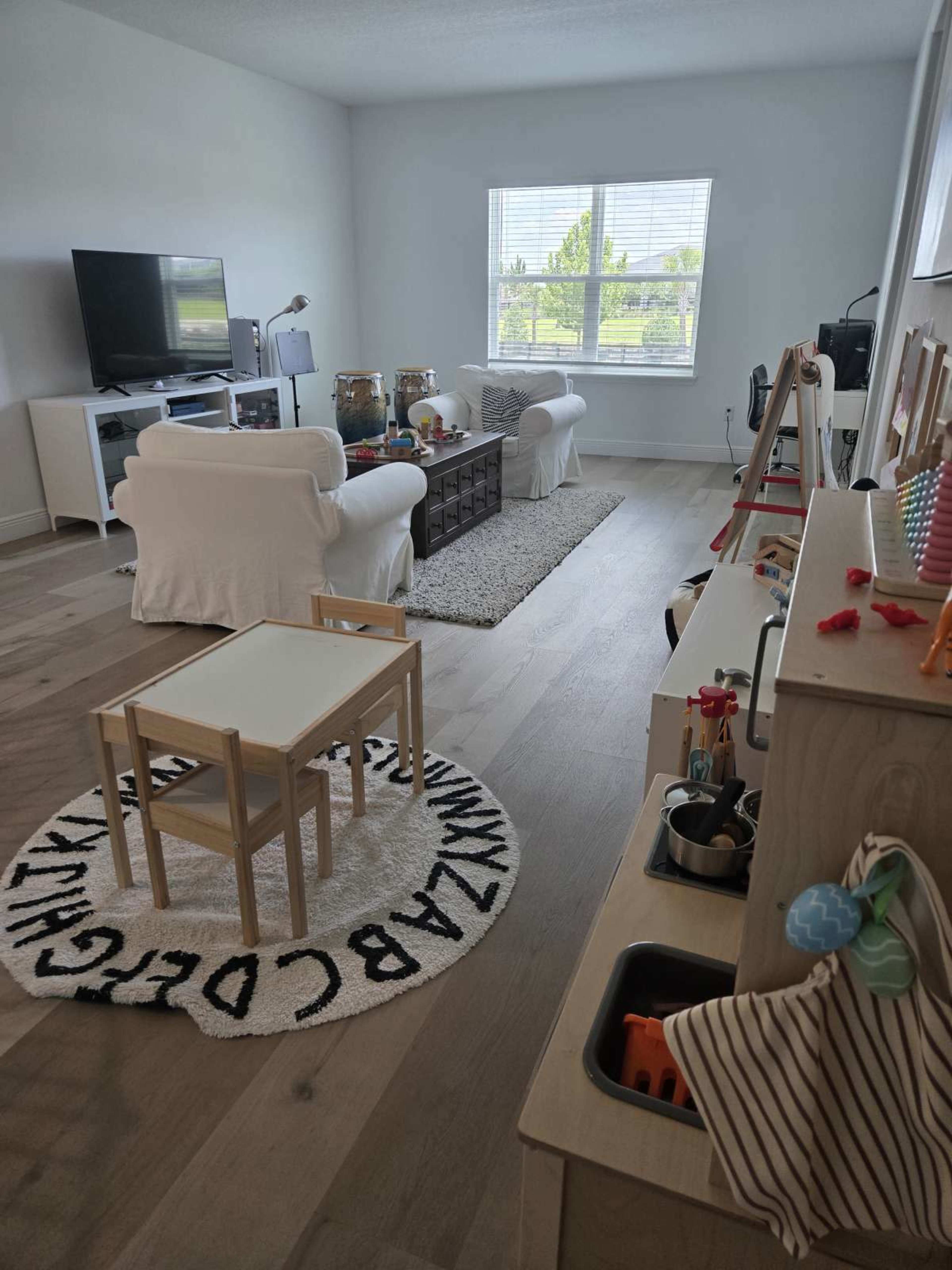 A bright living room features a seating area with white couches, a coffee table, and various children’s play items arranged near a window.