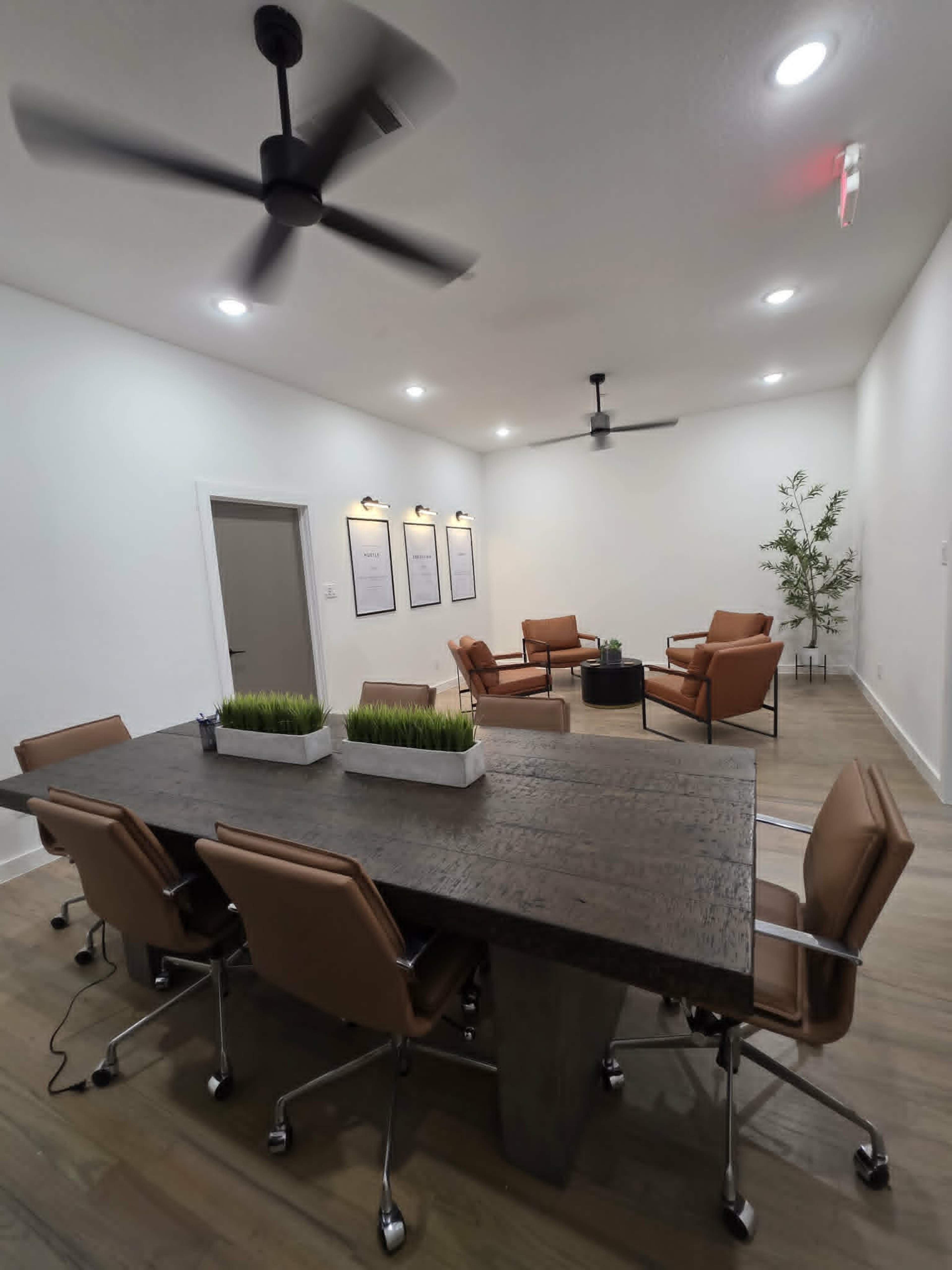 A modern office meeting room features a large wooden dining table surrounded by ergonomic chairs, with a seating area of leather chairs and a small table, all illuminated by overhead lights and ceiling fans.