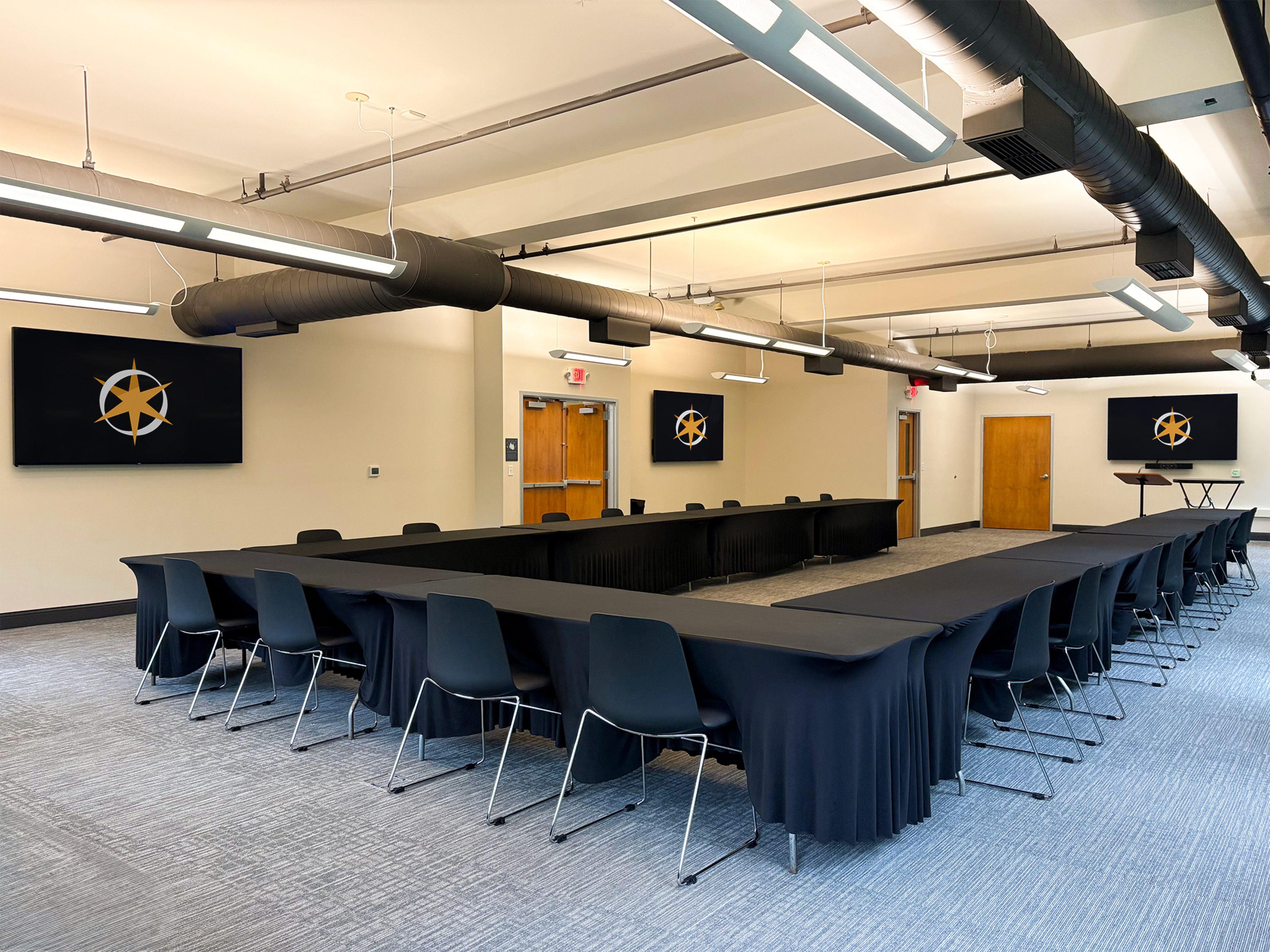 A conference room features a large rectangular table surrounded by black chairs, with two screens displaying a logo on the walls.