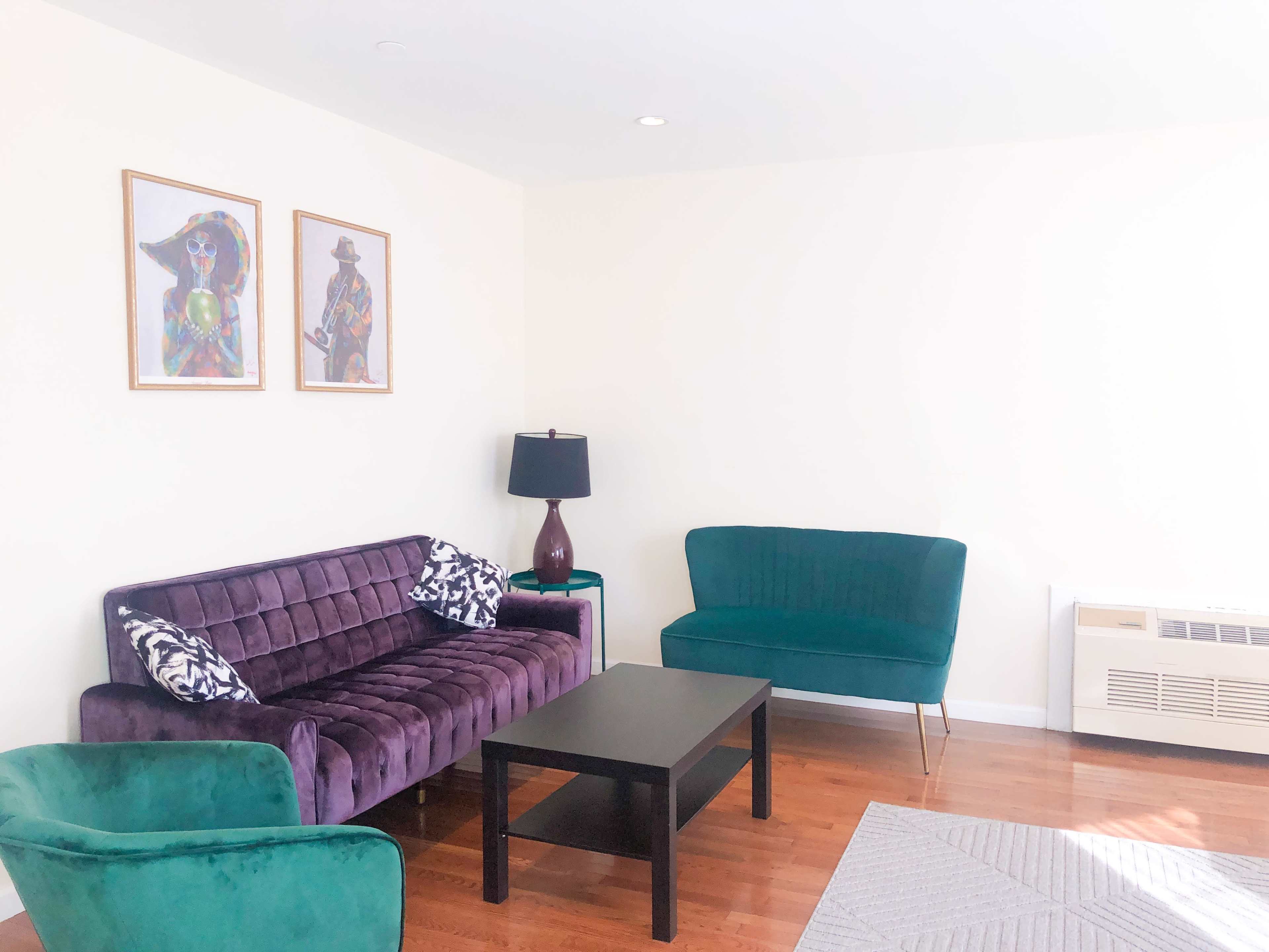 The image shows a living room with a purple sofa, a green cushioned chair, a teal loveseat, a black coffee table, and two framed artworks on the wall.