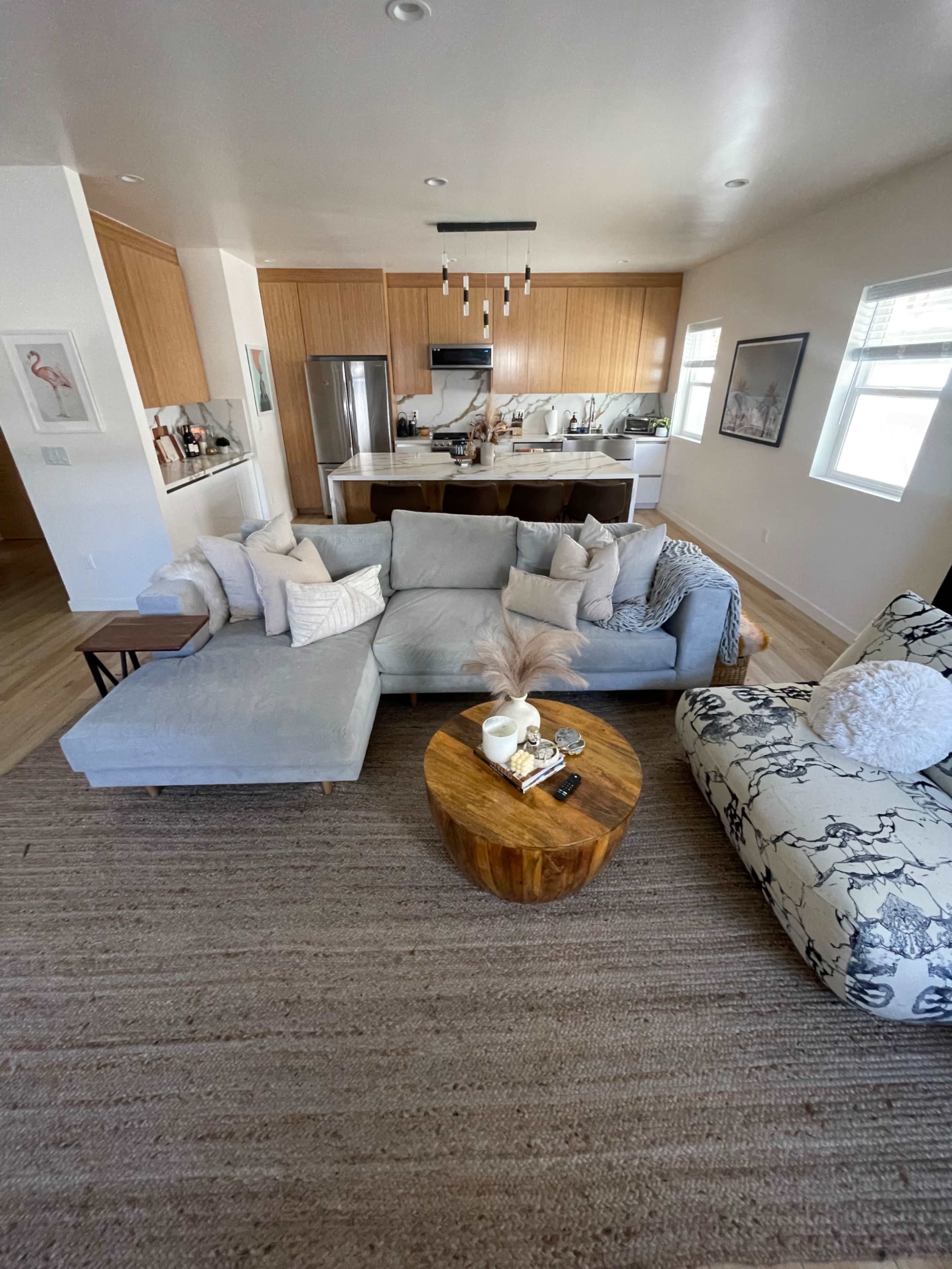 A living room features a sectional sofa, a round wooden coffee table, and a patterned armchair, with a view of an open kitchen in the background.