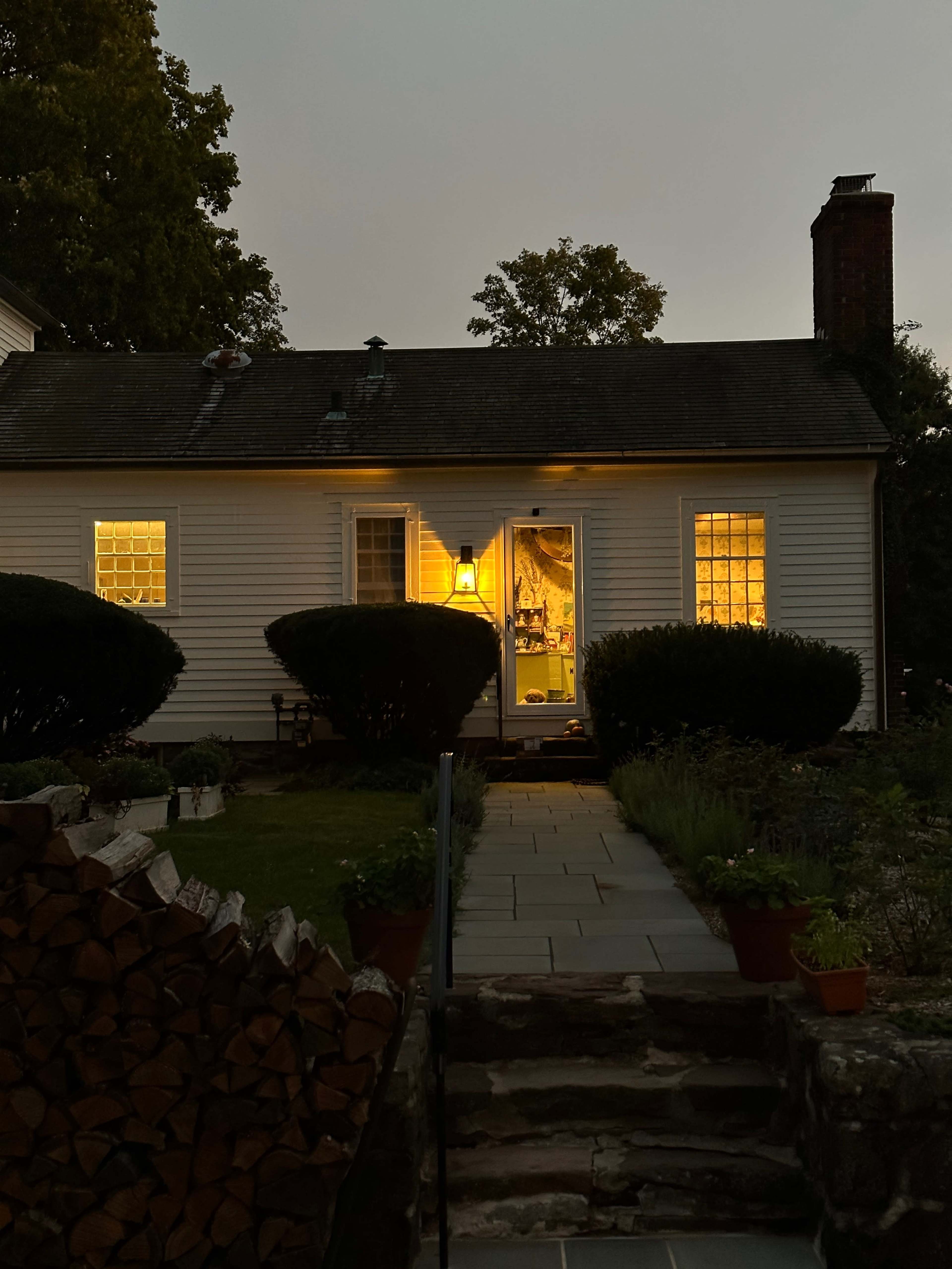 A well-lit farmhouse with glowing windows is set against a twilight sky, surrounded by greenery and neatly stacked firewood.