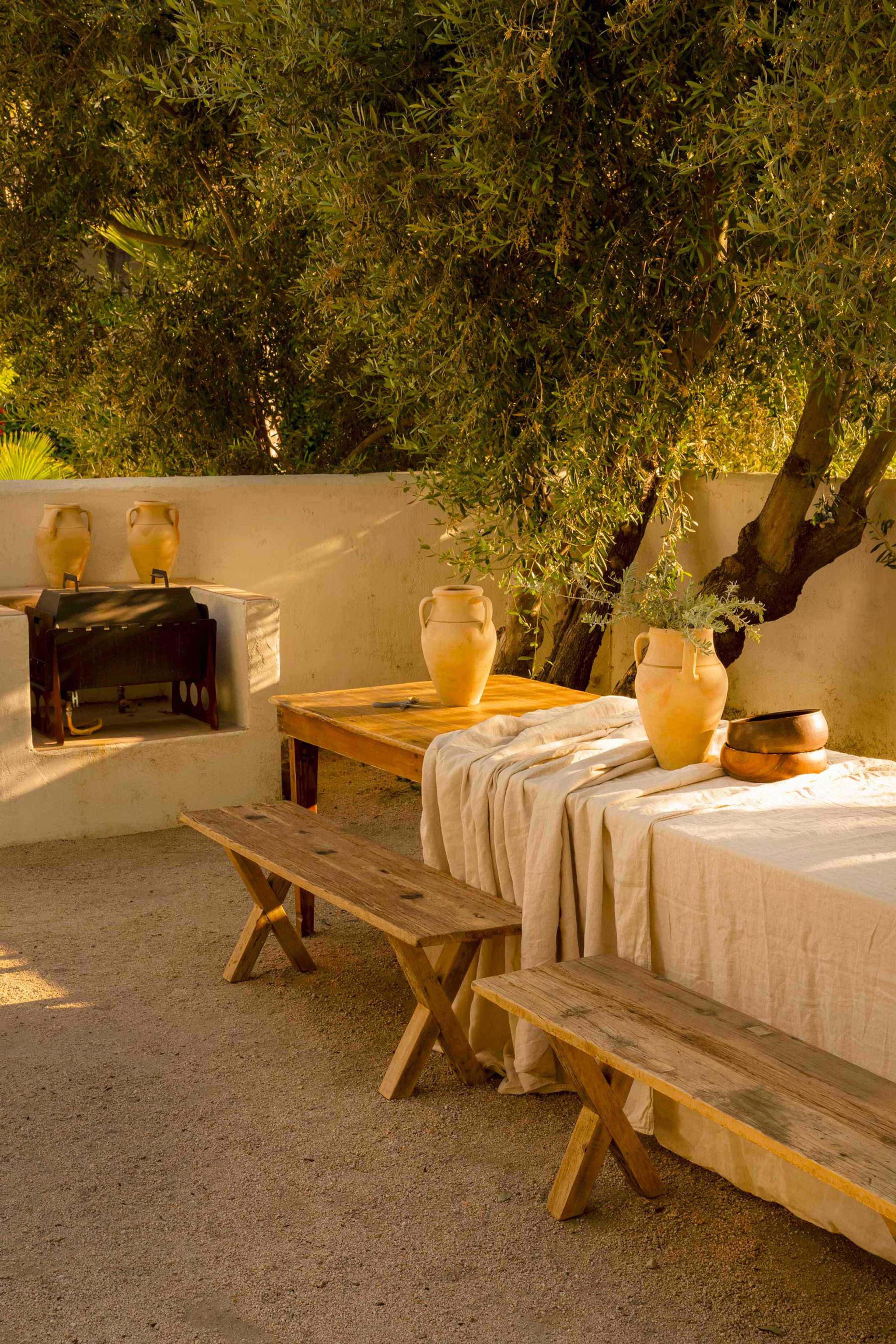A rustic outdoor dining area features wooden tables and benches, with clay pots displayed on a table beneath an olive tree.