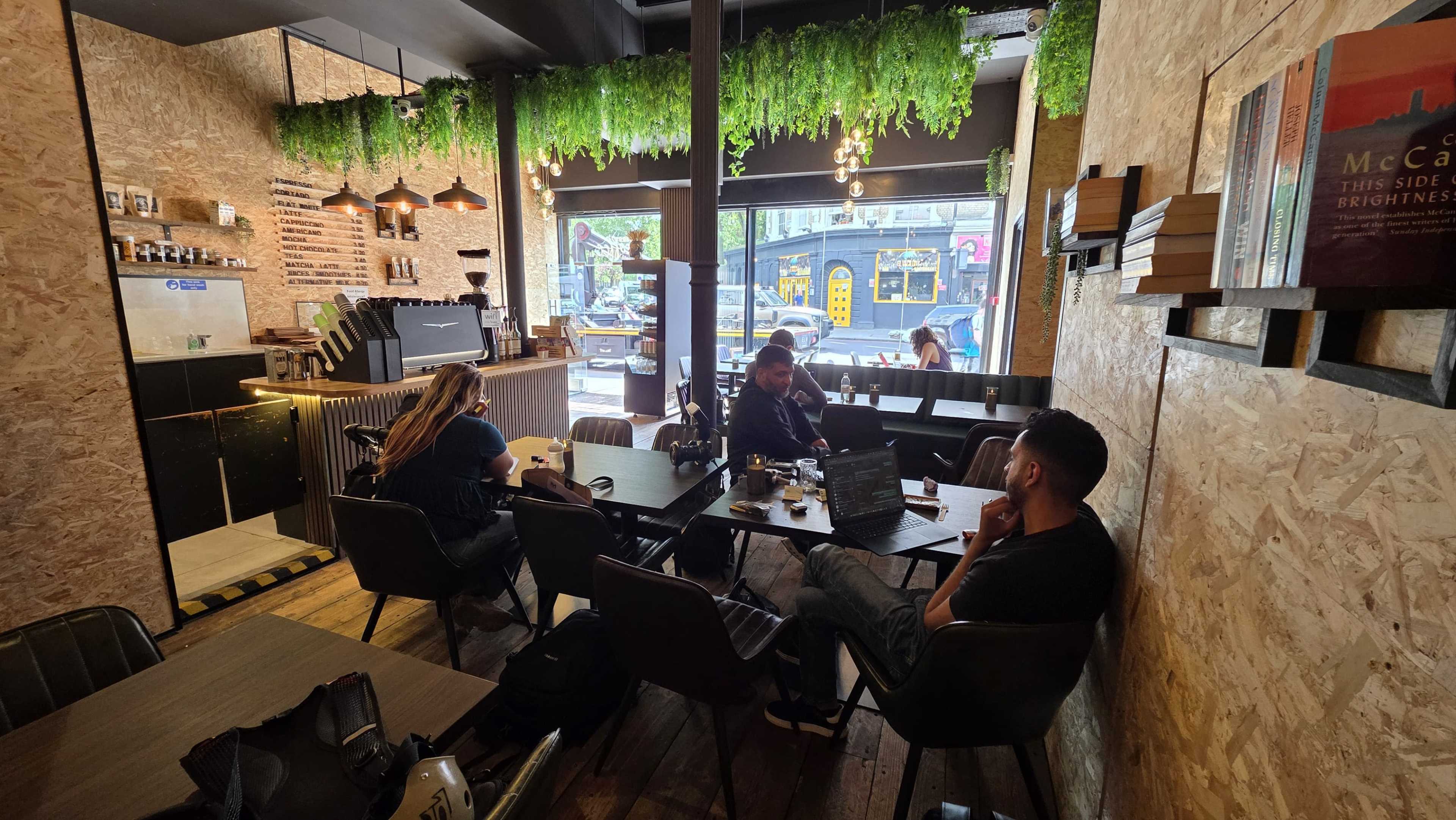 A cozy café interior features wooden furnishings and hanging greenery, with patrons working on laptops at tables near a large window.