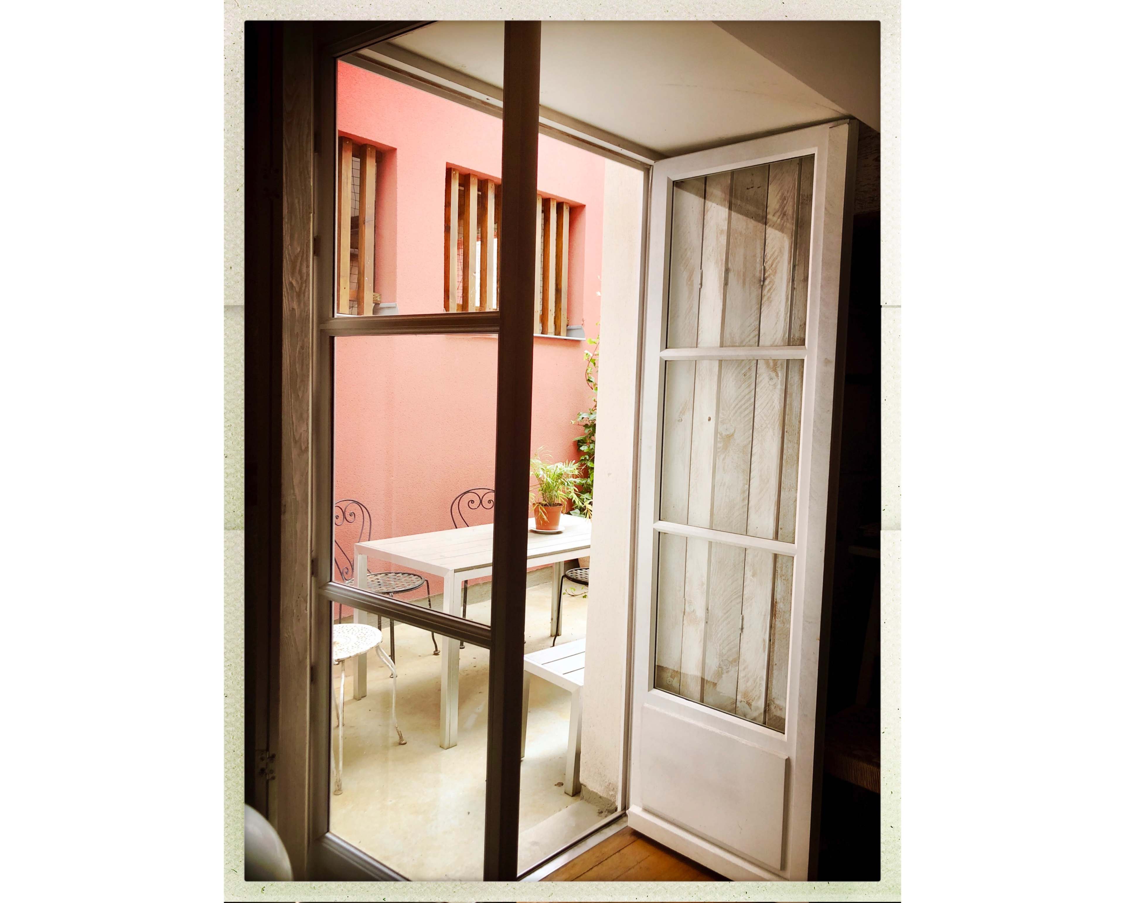 A white framed door opens to a small outdoor space with a table and chairs against a pink wall.
