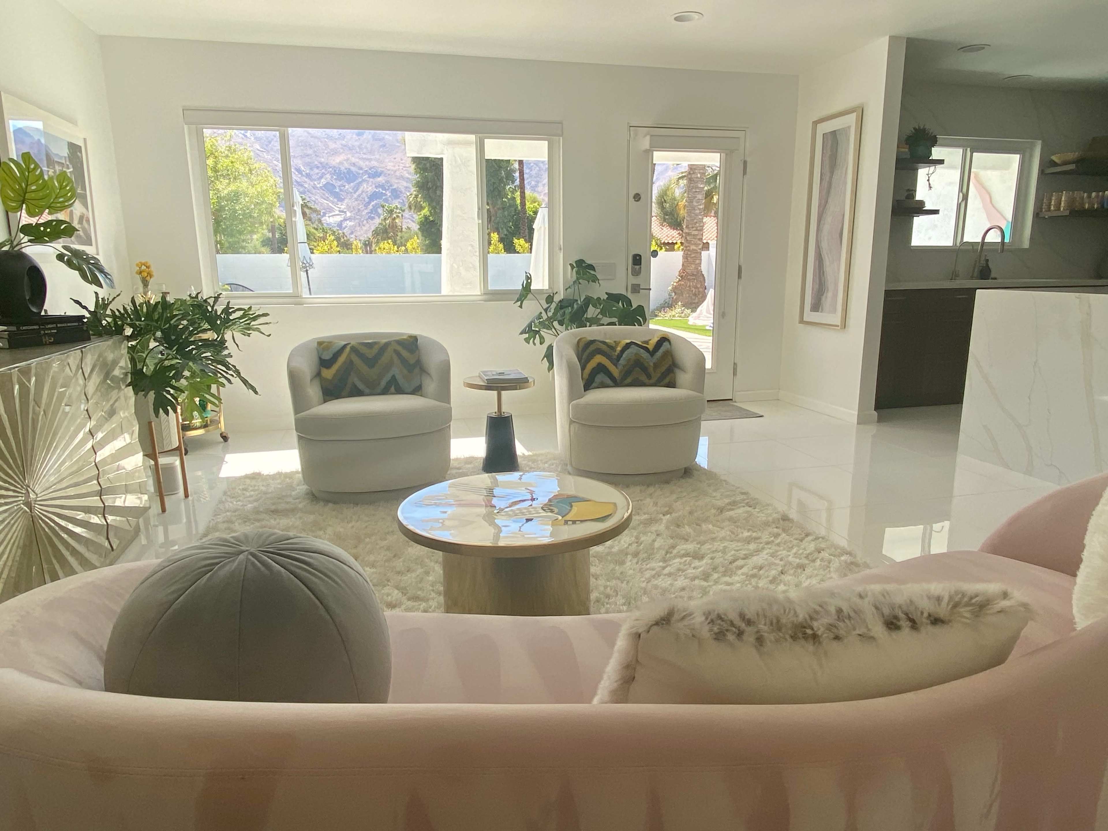 A modern living room with two white chairs and a pink sofa, a round coffee table, and large windows that provide natural light and a view of palm trees and mountains.