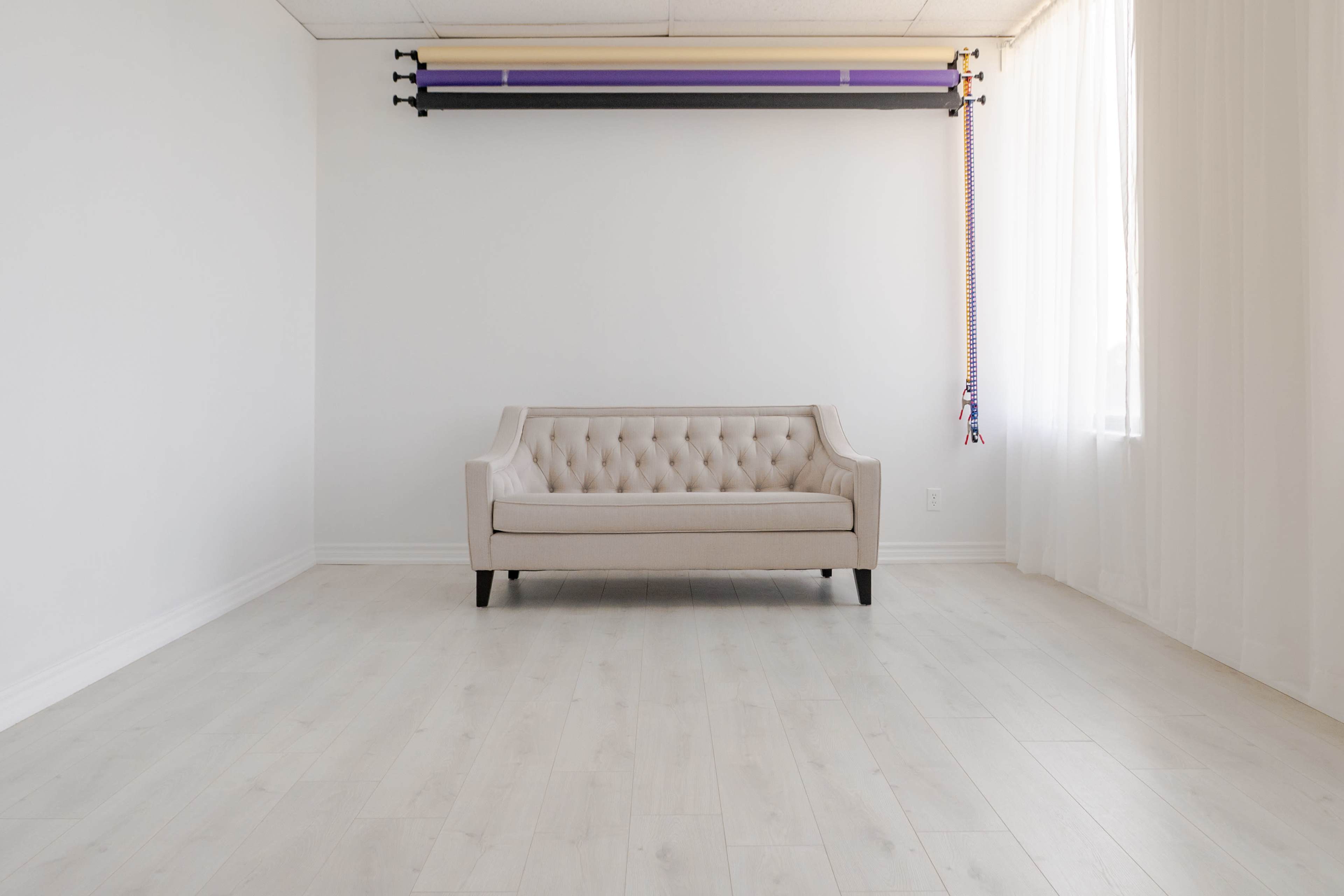 A light-colored, tufted sofa is positioned in the center of a spacious, empty room with white walls and a large window.