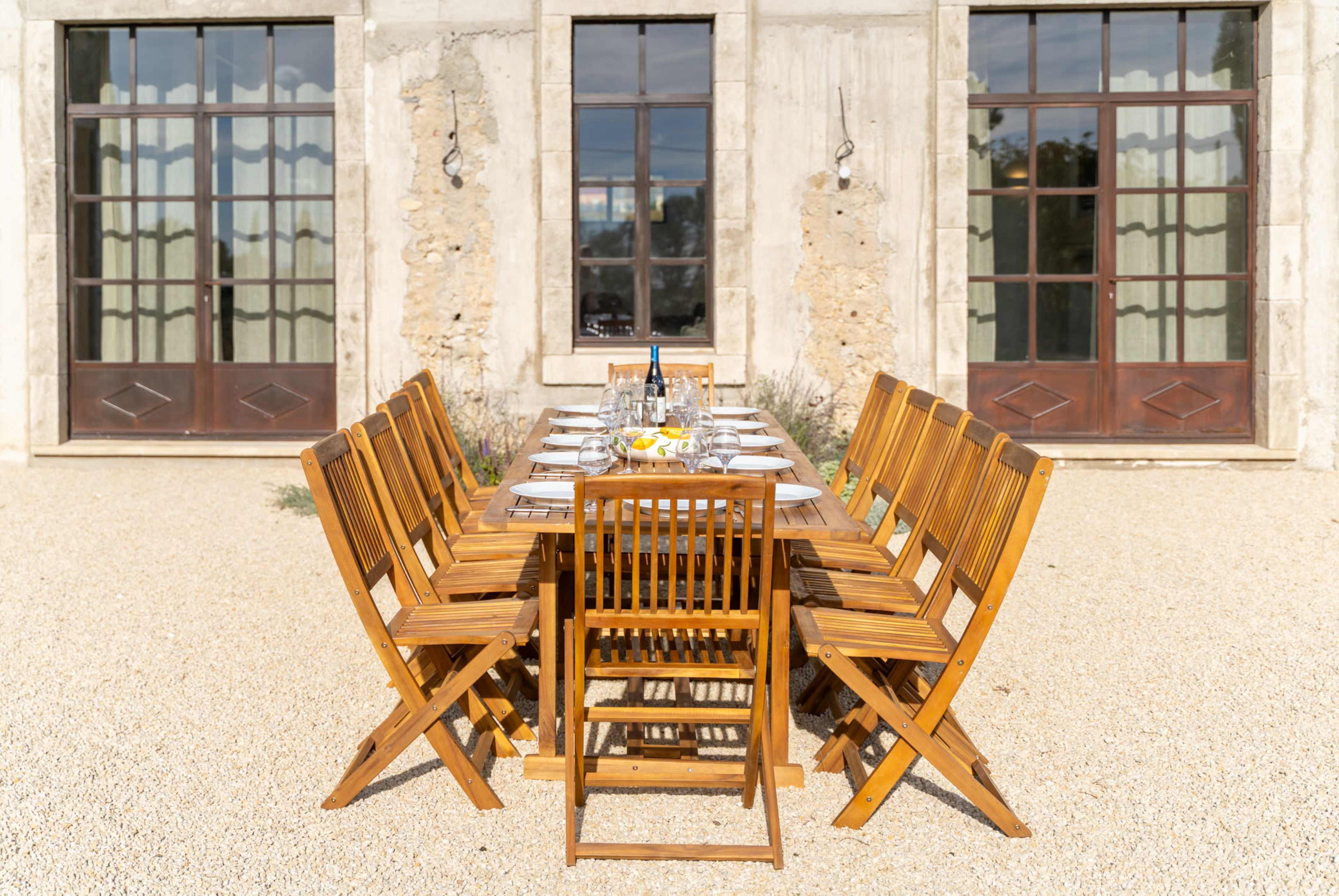 A long wooden dining table is set with dishes and seating for guests, placed outside a building with large windows and doors.