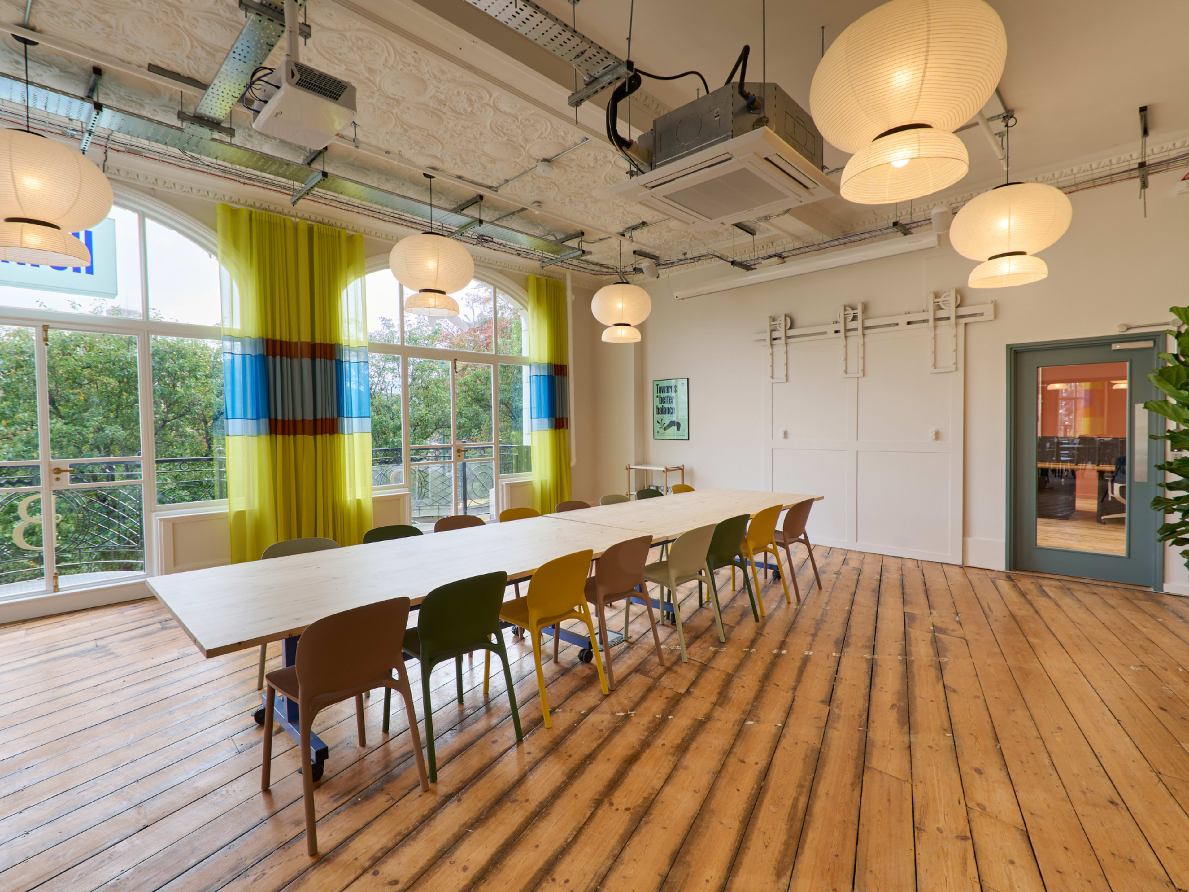 A large meeting room features a long wooden table surrounded by colorful chairs, with large windows allowing natural light to fill the space.