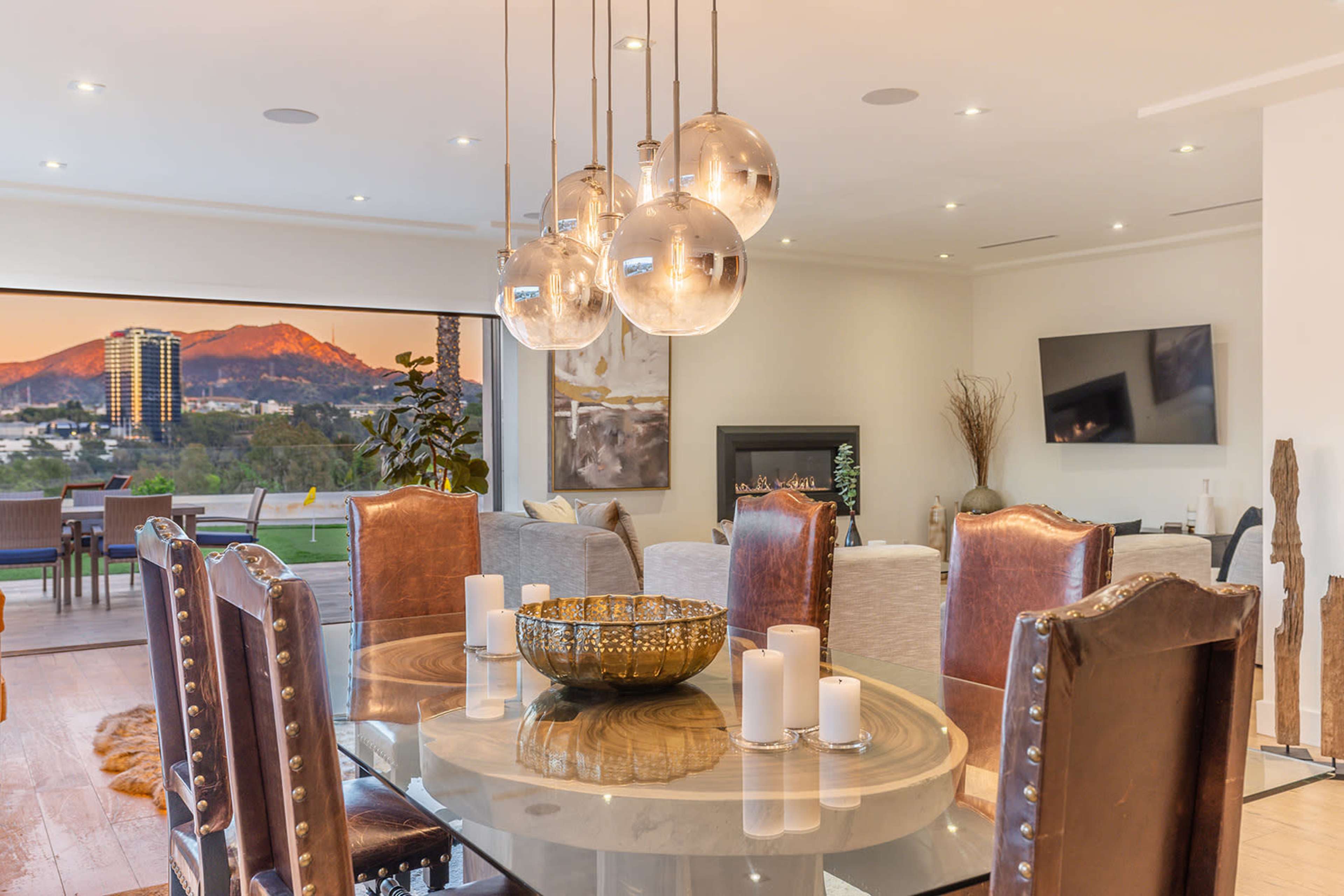 A dining area features a round glass table surrounded by upholstered chairs, with a backdrop of large windows showing a mountain view and pendant lighting above.