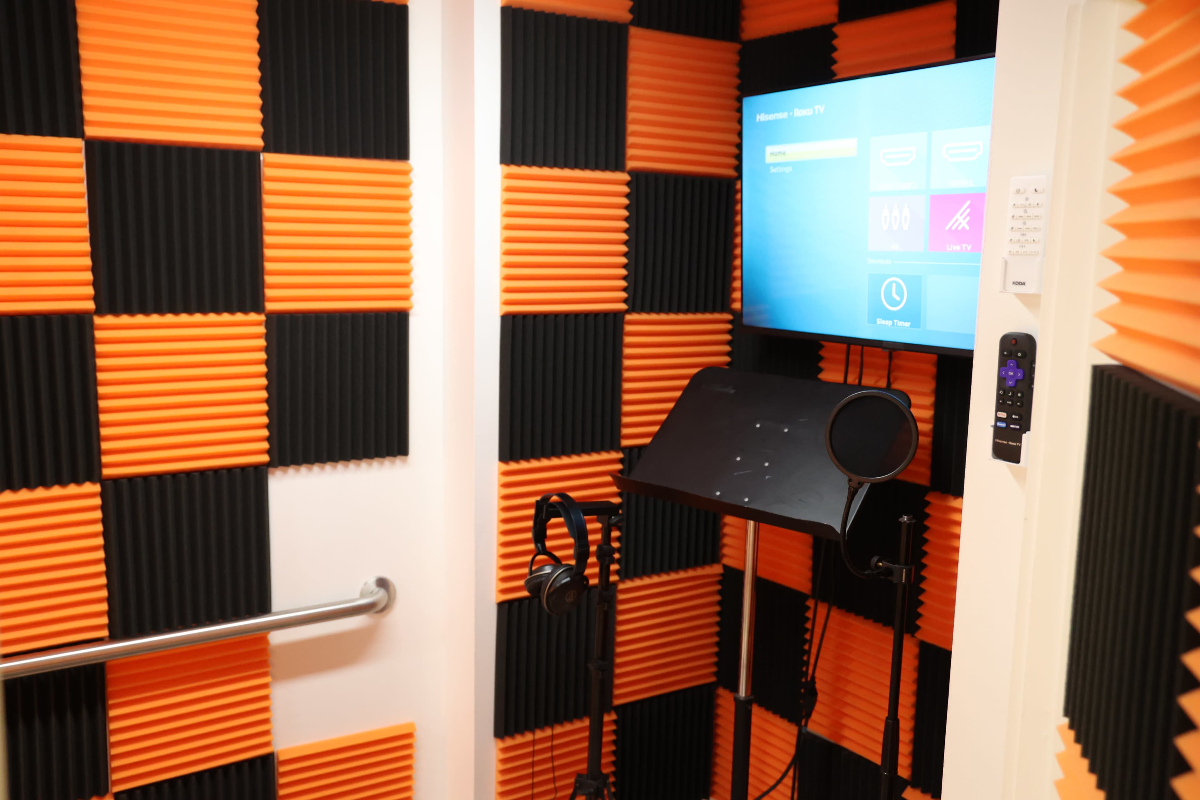 The image shows a soundproof recording studio with orange and black acoustic foam panels on the walls, a monitor displaying options, and a microphone setup.