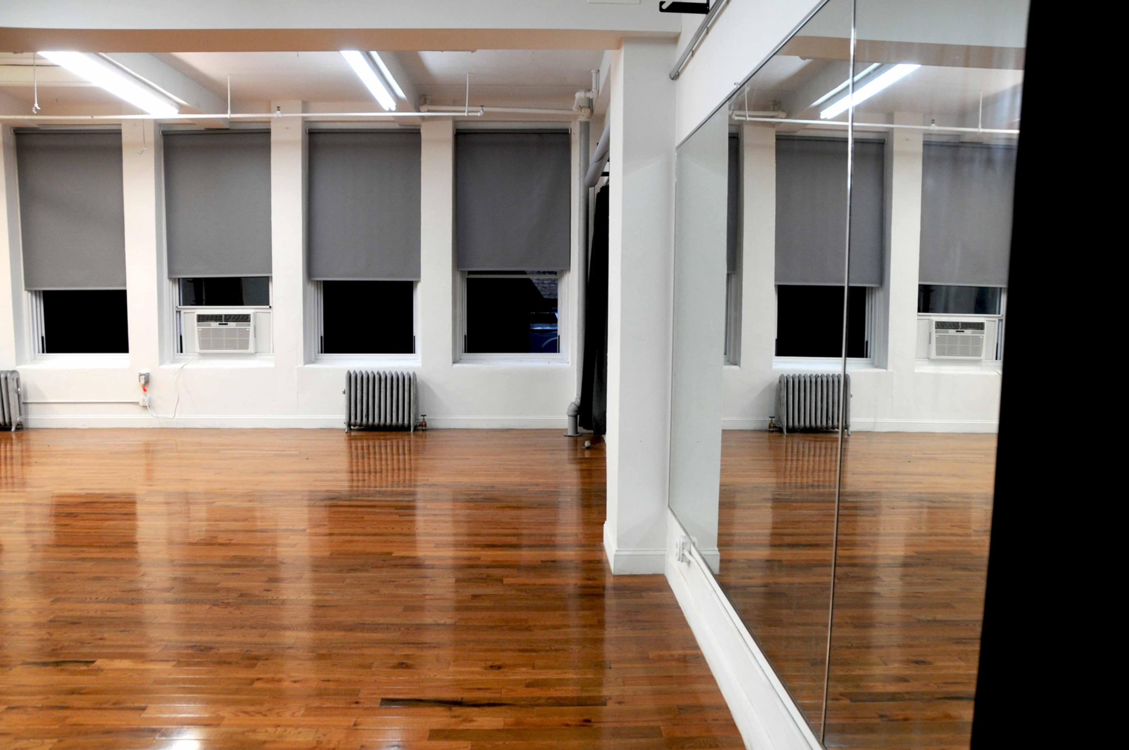 A spacious interior room with hardwood floors, large windows covered with gray shades, and a mirrored wall reflecting the empty space.