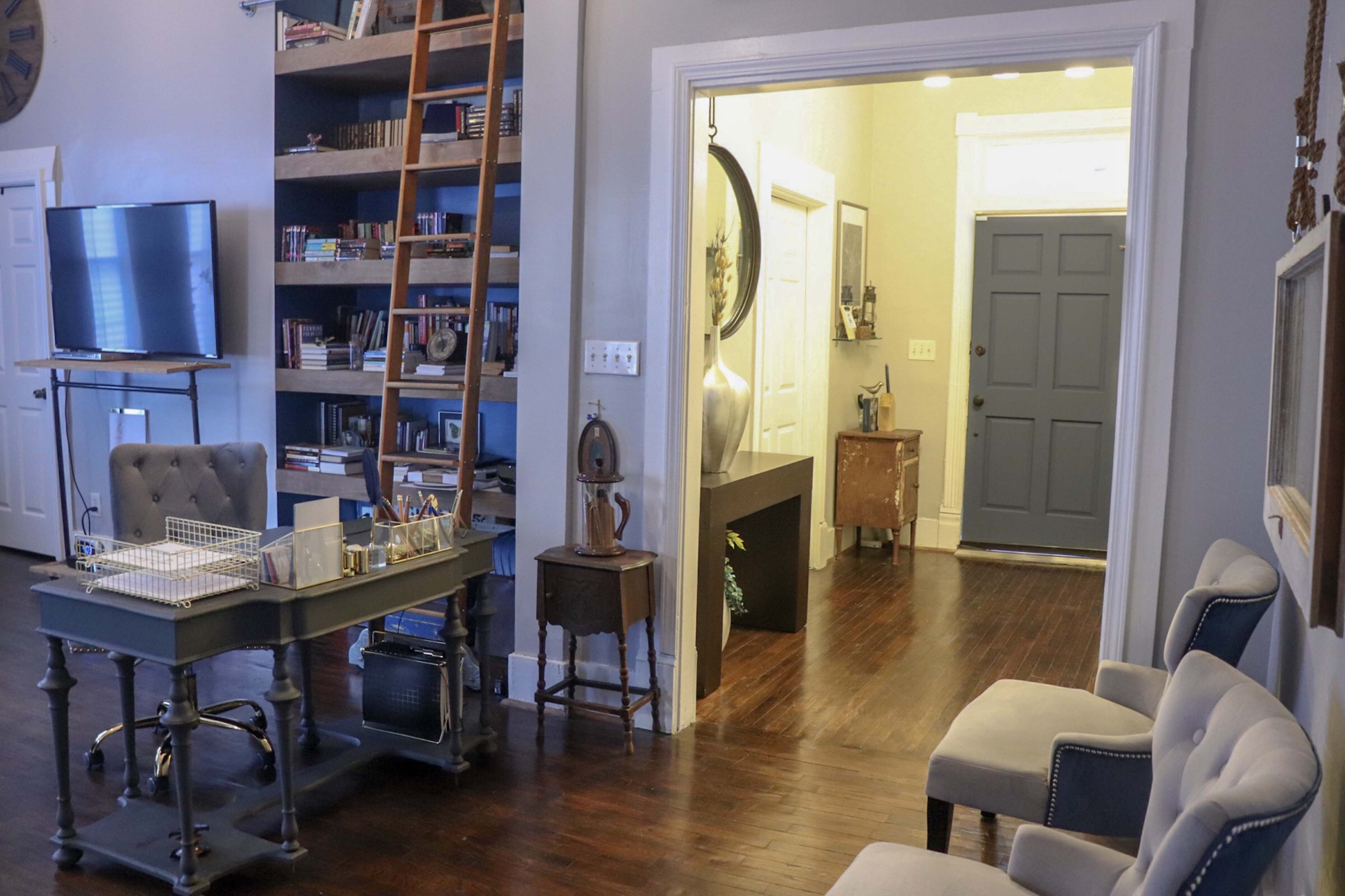A cozy interior space with a desk on the left, a ladder bookshelf, and a hallway leading to a door in the background.