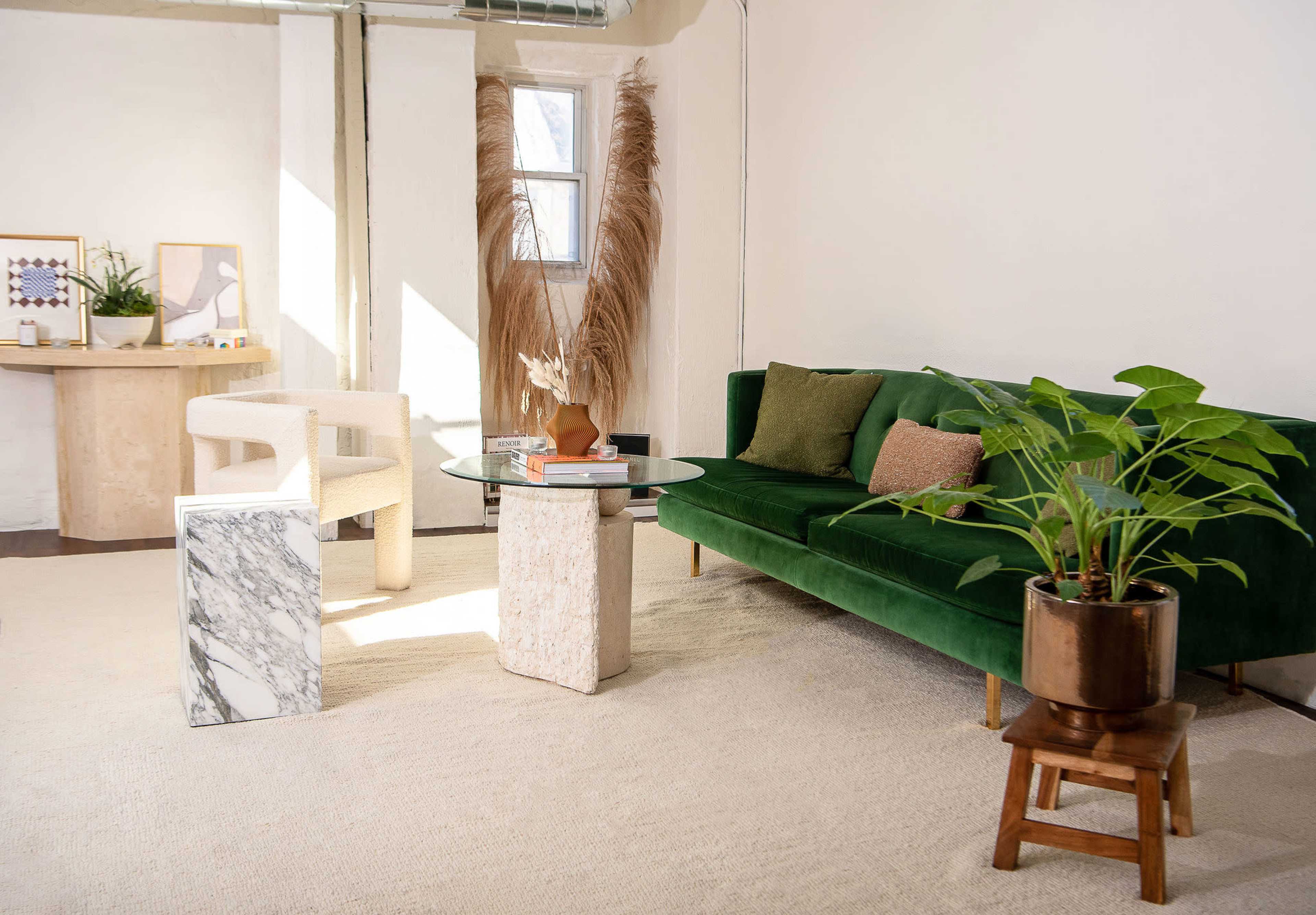 A modern living space with a green sofa, a glass coffee table placed on a marble and stone base, and a potted plant near a light-colored rug.