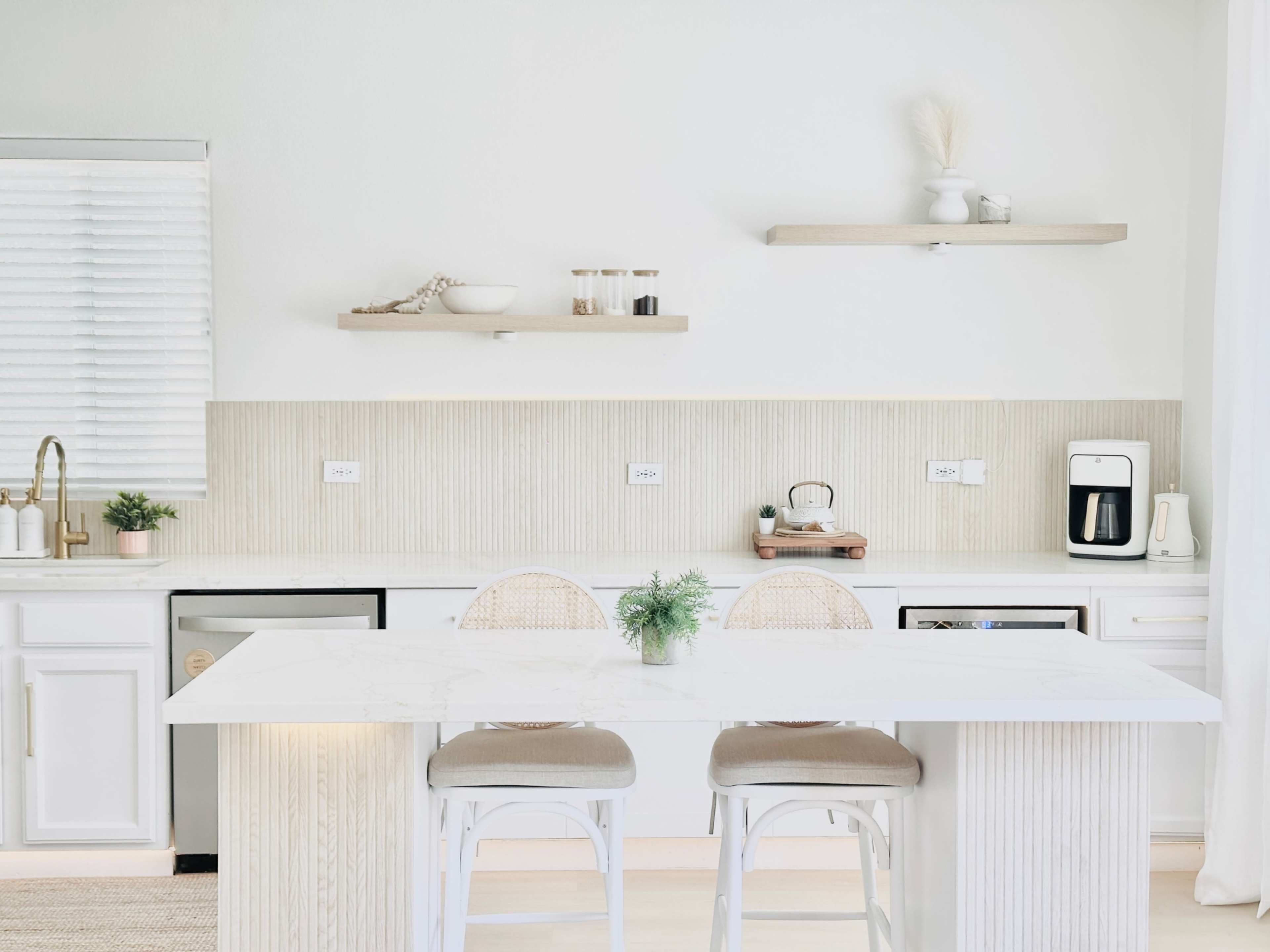 A modern kitchen features a marble island, white cabinetry, and wooden shelves adorned with decorative items.
