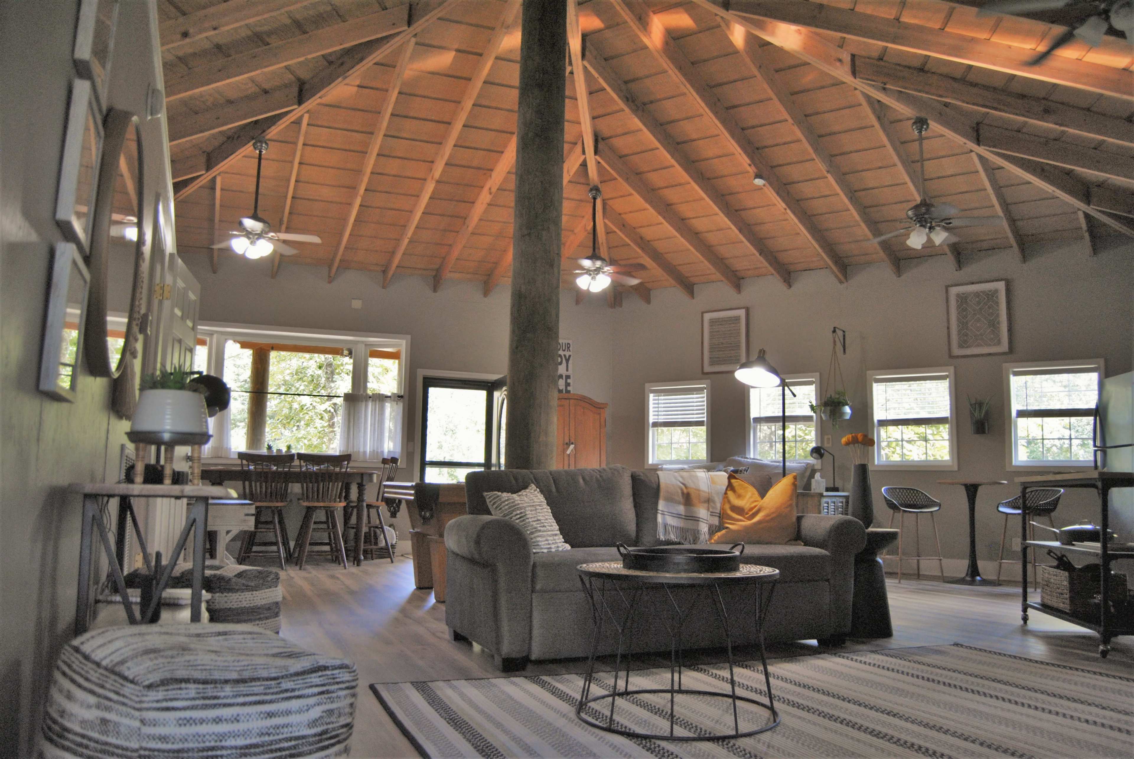 An open-concept living space with a high, wooden vaulted ceiling, featuring a gray sofa, a coffee table, and a dining area with a large window.