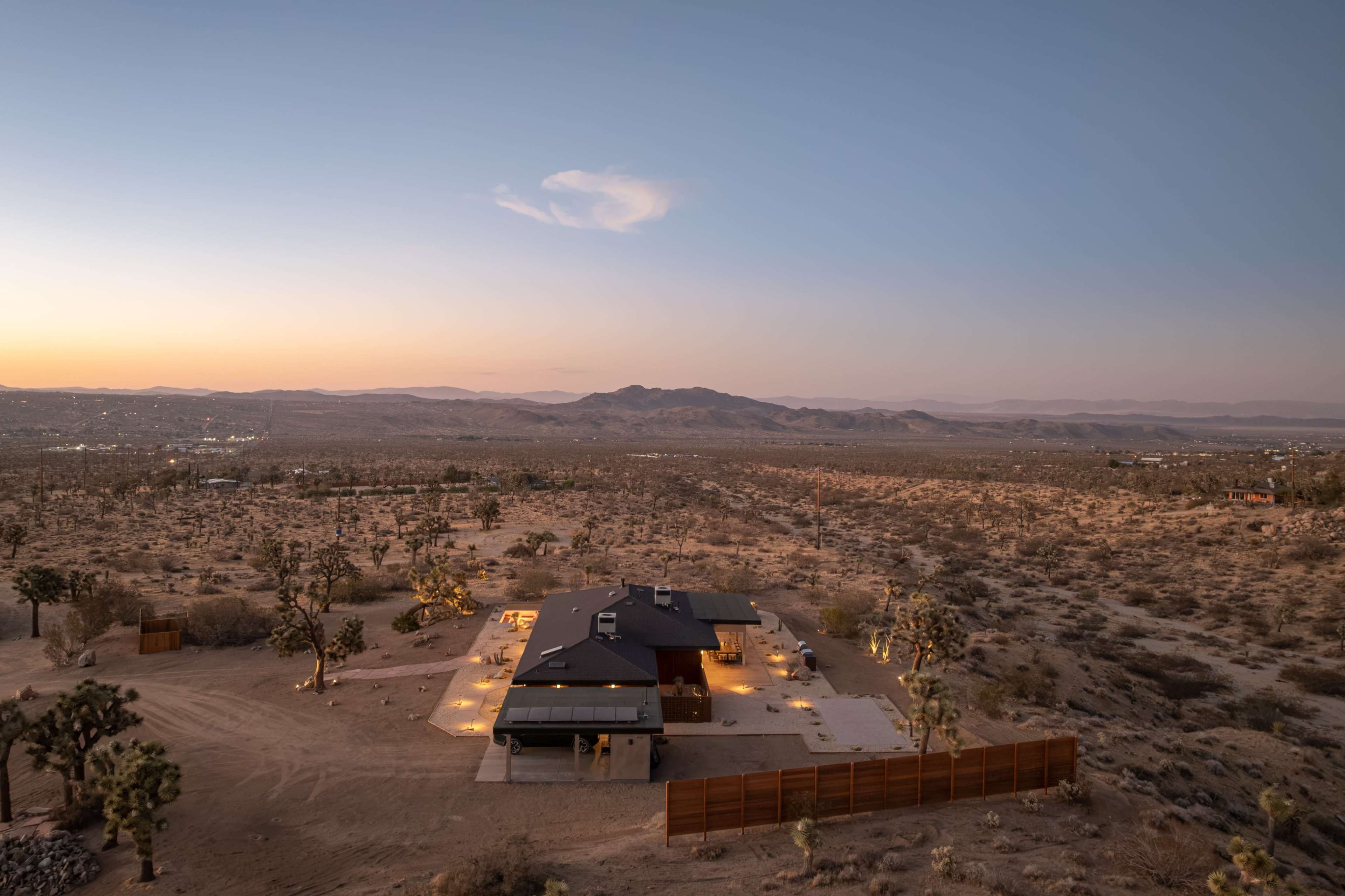 A modern house sits in a desert landscape at dusk, surrounded by sparse vegetation and mountains in the distance.