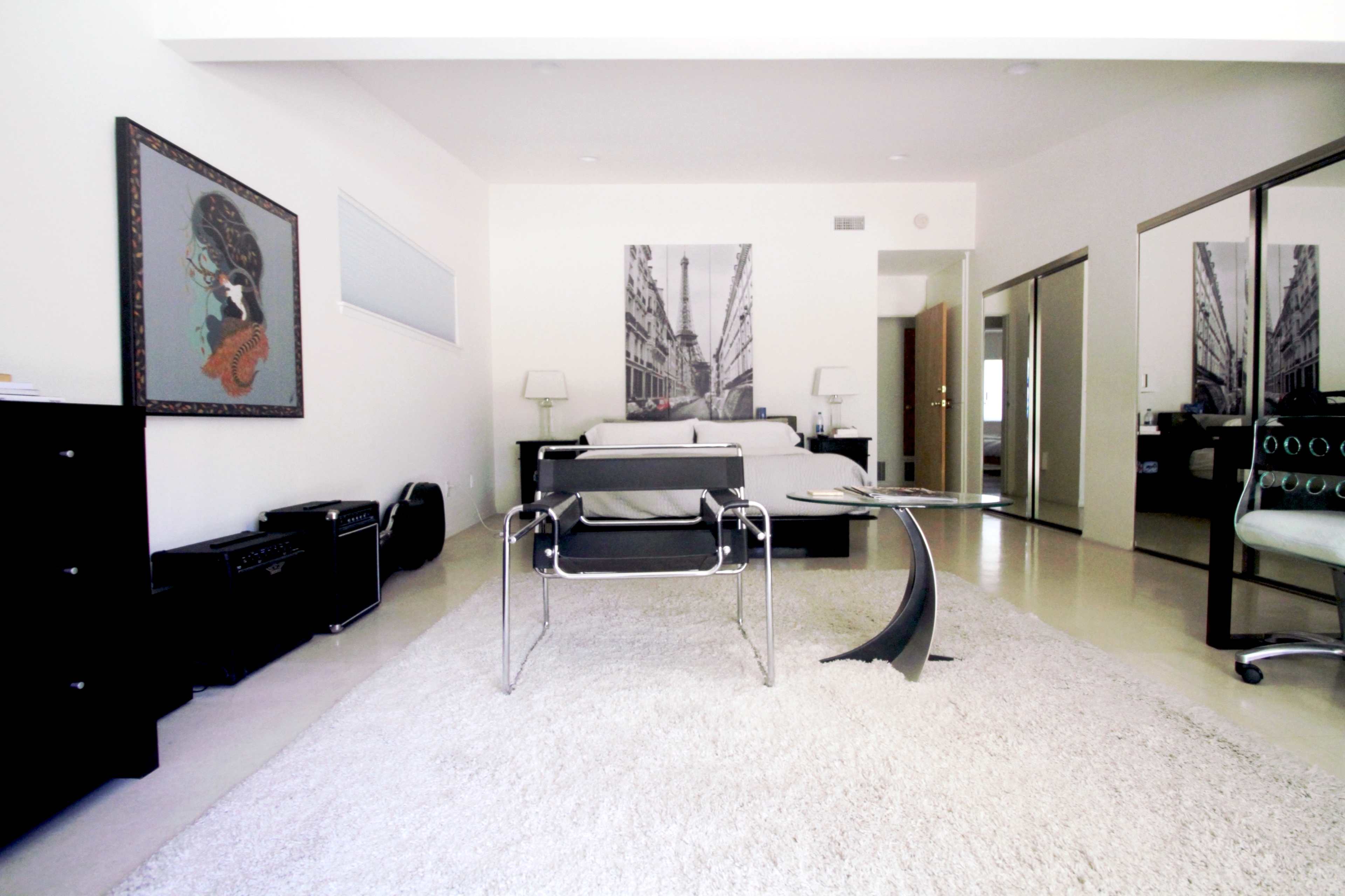 A modern living space with a bed, large mirror, and a small coffee table on a carpeted floor, complemented by artwork on the walls.