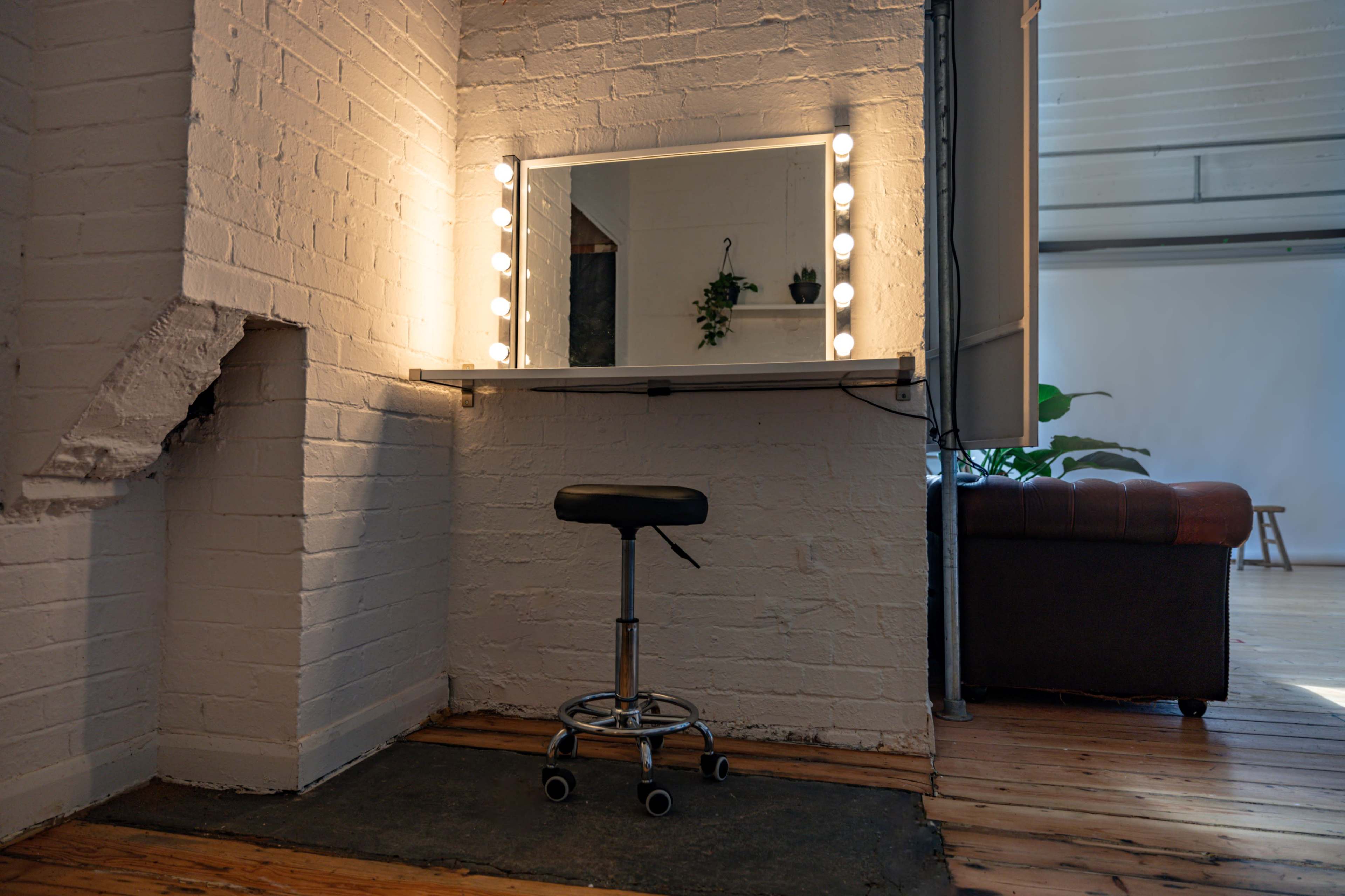 A well-lit vanity mirror with a stool is positioned against a white brick wall in a room with wooden flooring.