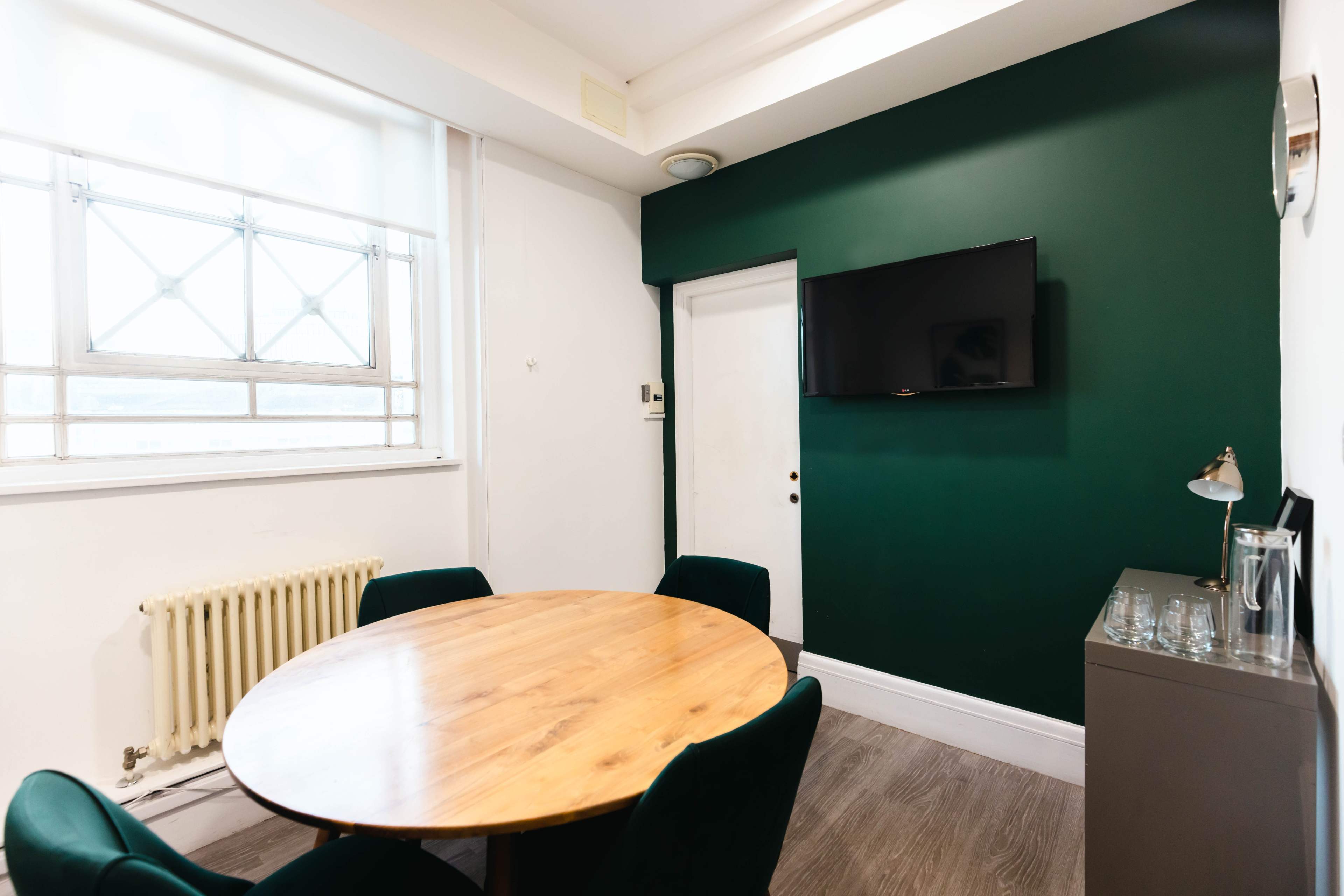 The image shows a small meeting room with a round wooden table, four green chairs, a wall-mounted television, and a radiator beside a window.