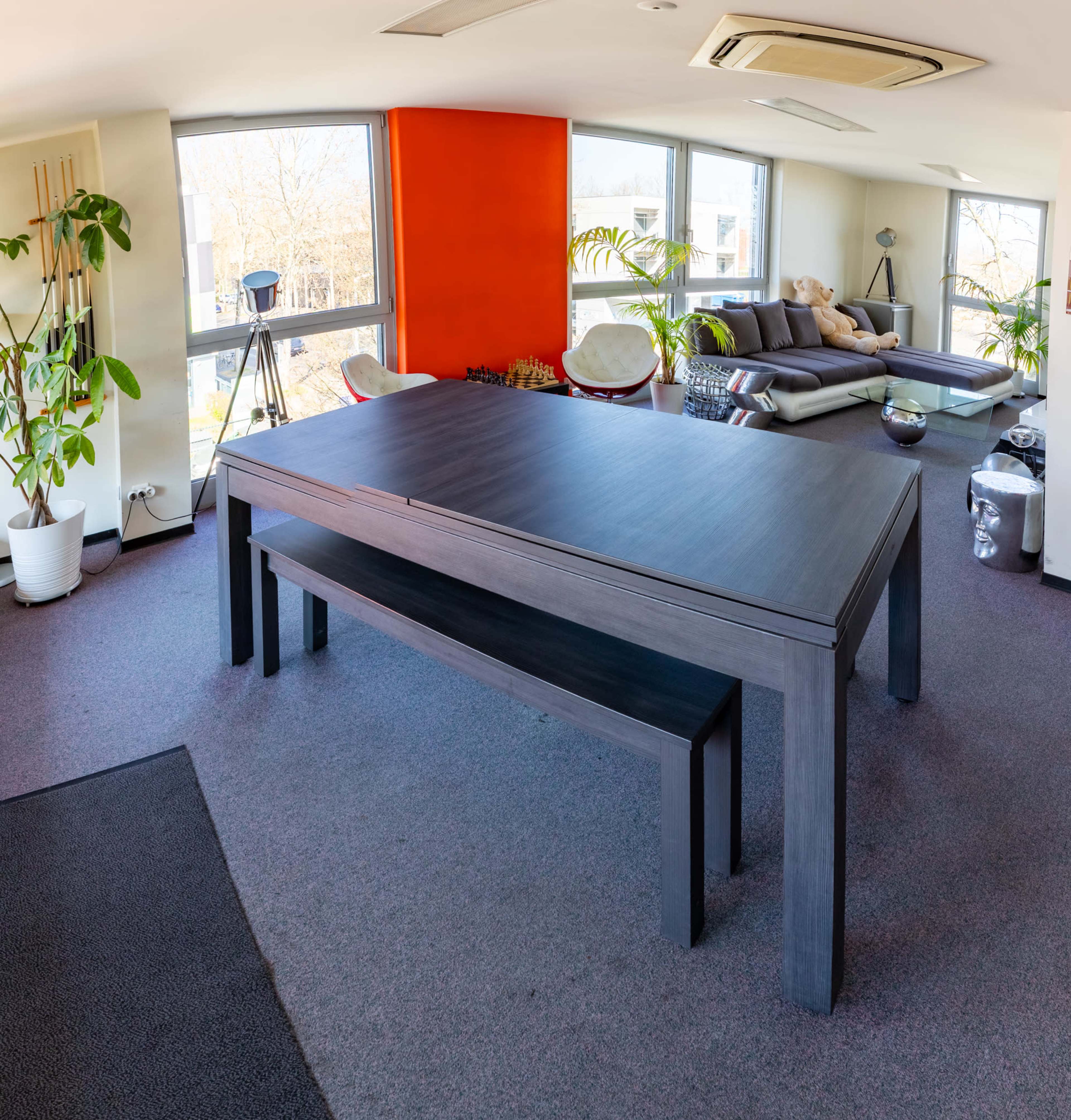 A spacious room features a large dark wooden table with a matching bench, surrounded by plants and a modern seating area.