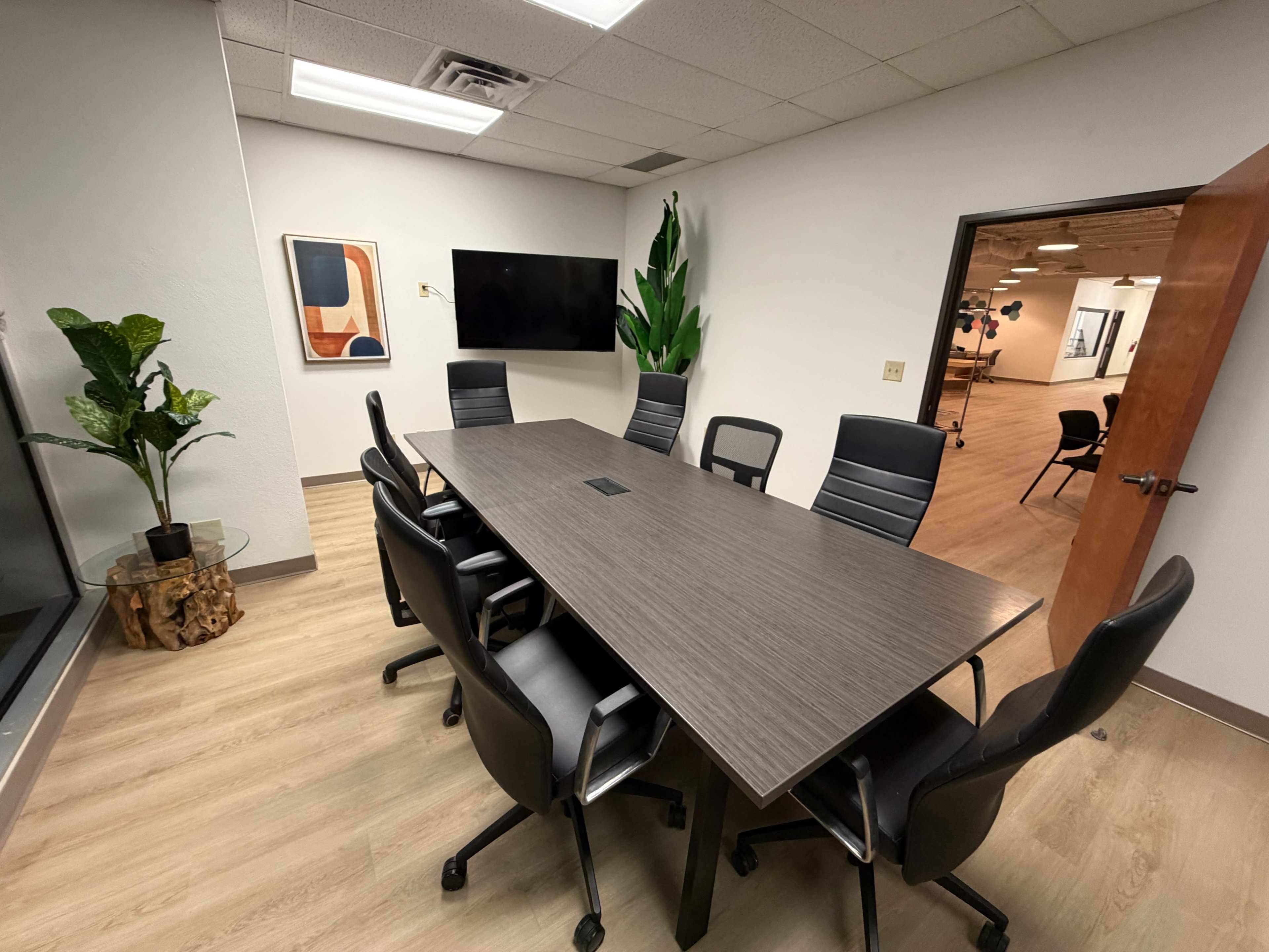 The image shows a modern conference room with a long table surrounded by black office chairs, a wall-mounted screen, and a large plant in the corner.