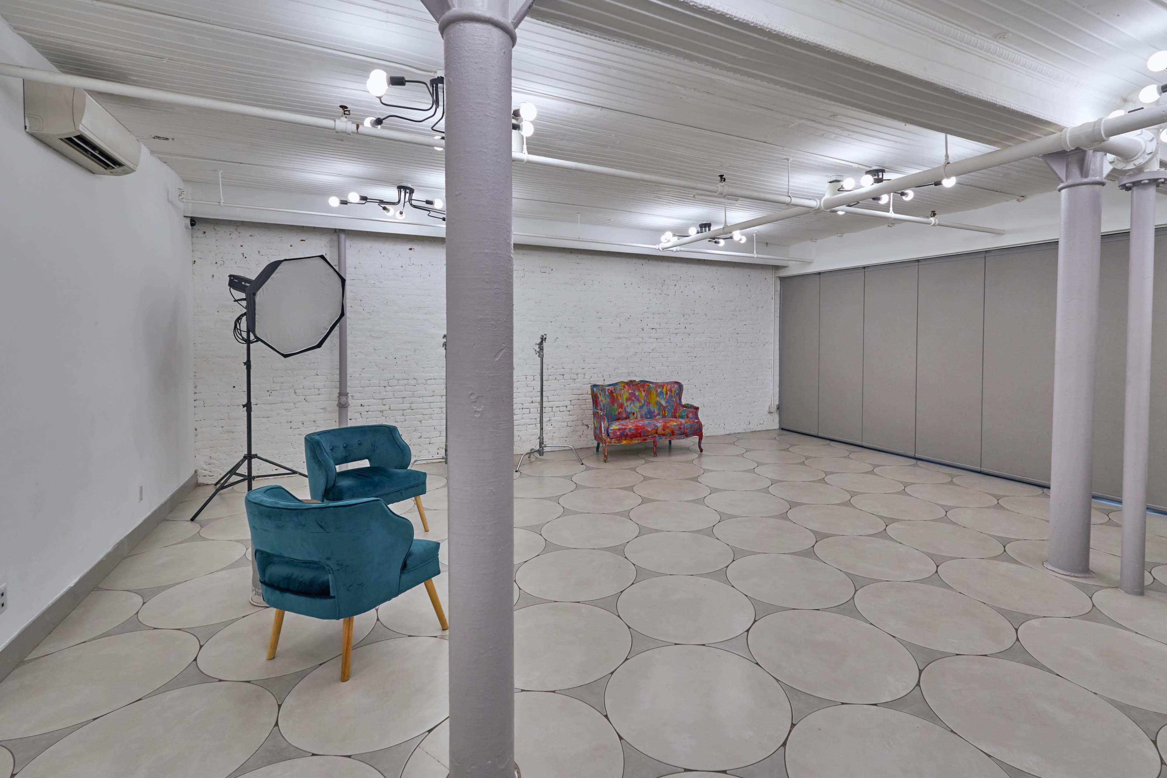 A spacious room with a patterned tile floor, two chairs, a photography light setup, and movable partition panels against a white brick wall.