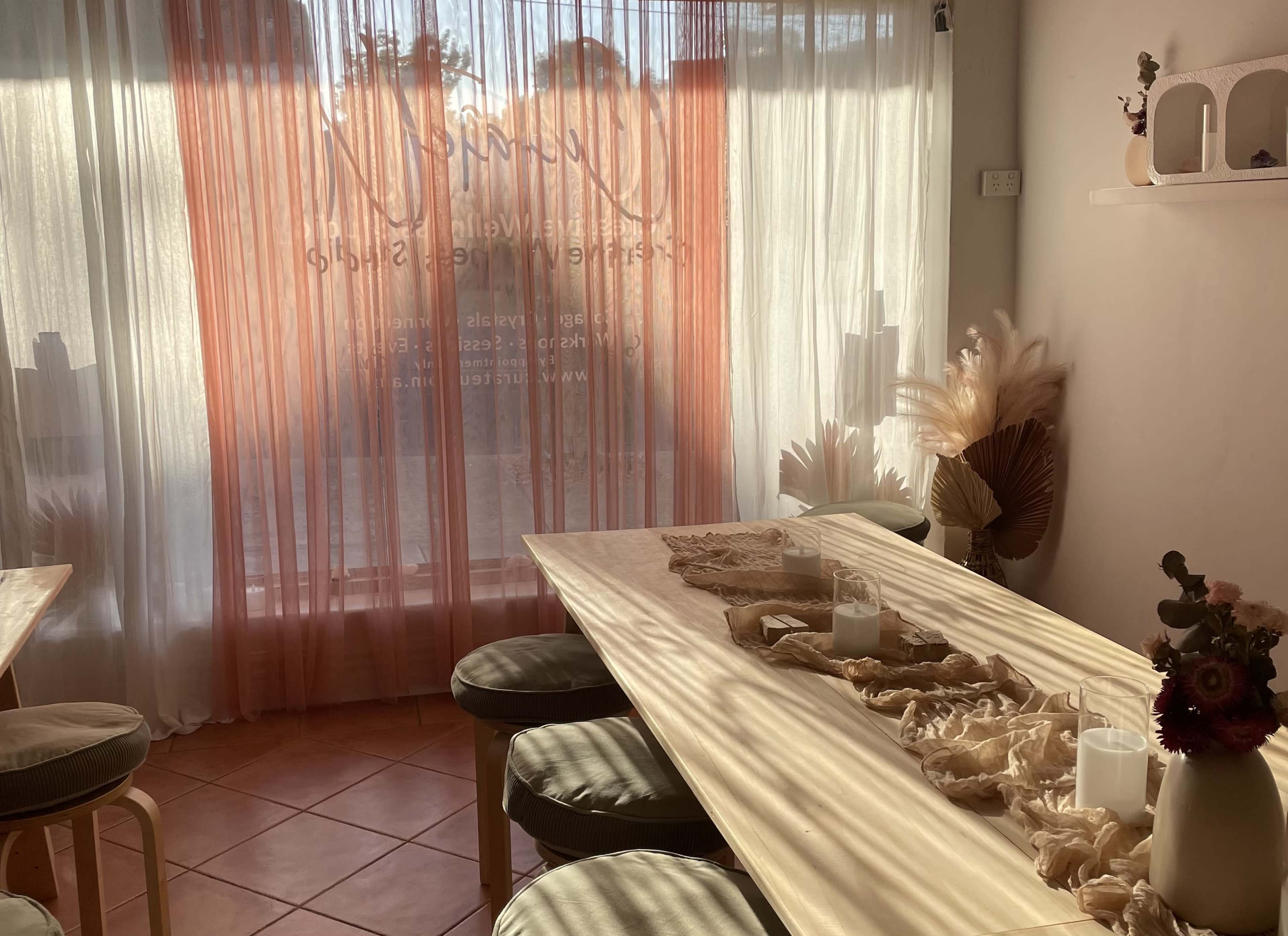 The image shows a softly lit, minimalist interior of a café or workspace with wooden tables, cushions, and sheer curtains.
