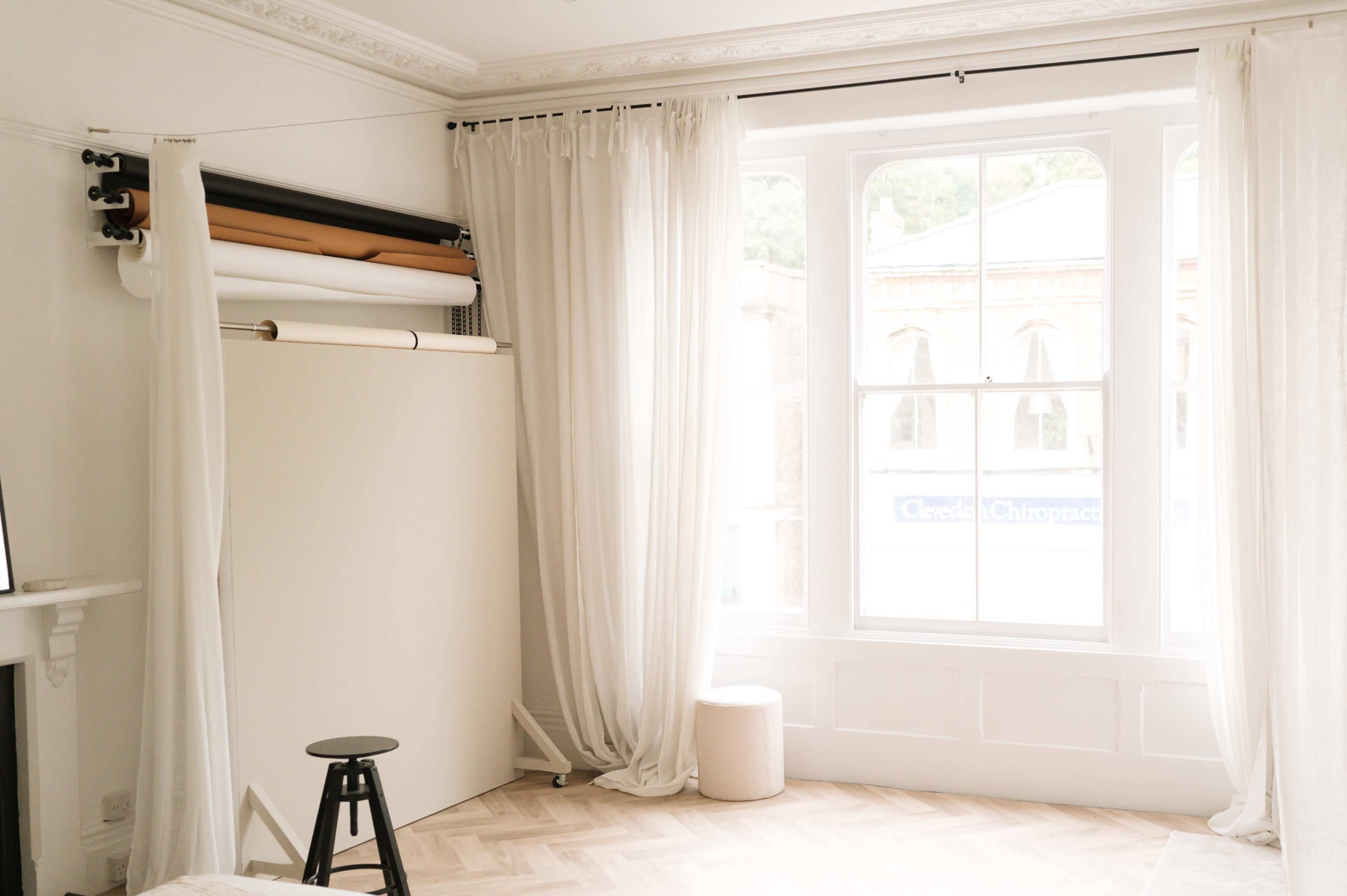 A minimalist interior with a white backdrop, a stool, and large windows adorned with sheer curtains.