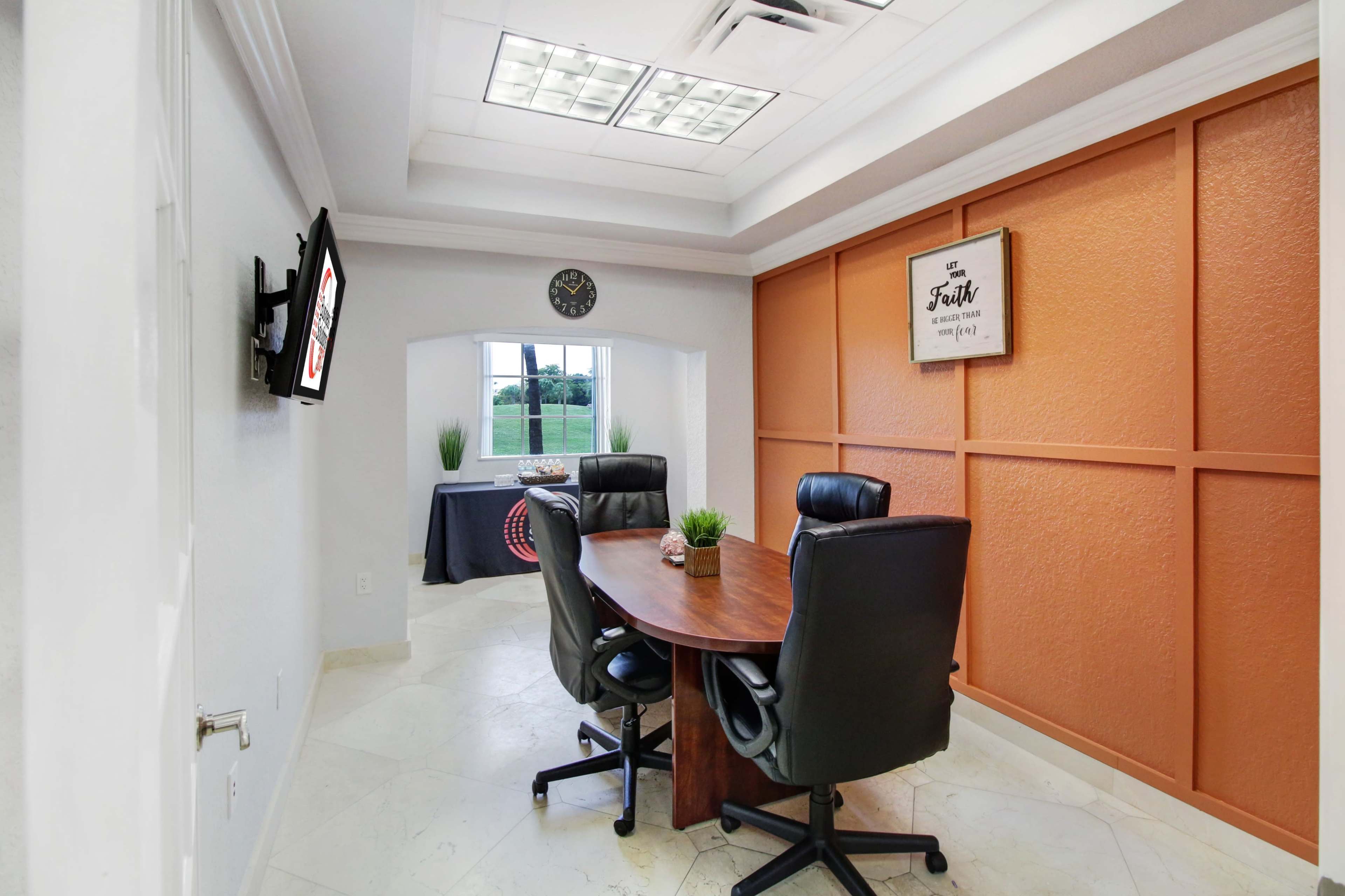 The image shows a small conference room with a round wooden table, four black leather chairs, and a large window overlooking a grassy area.
