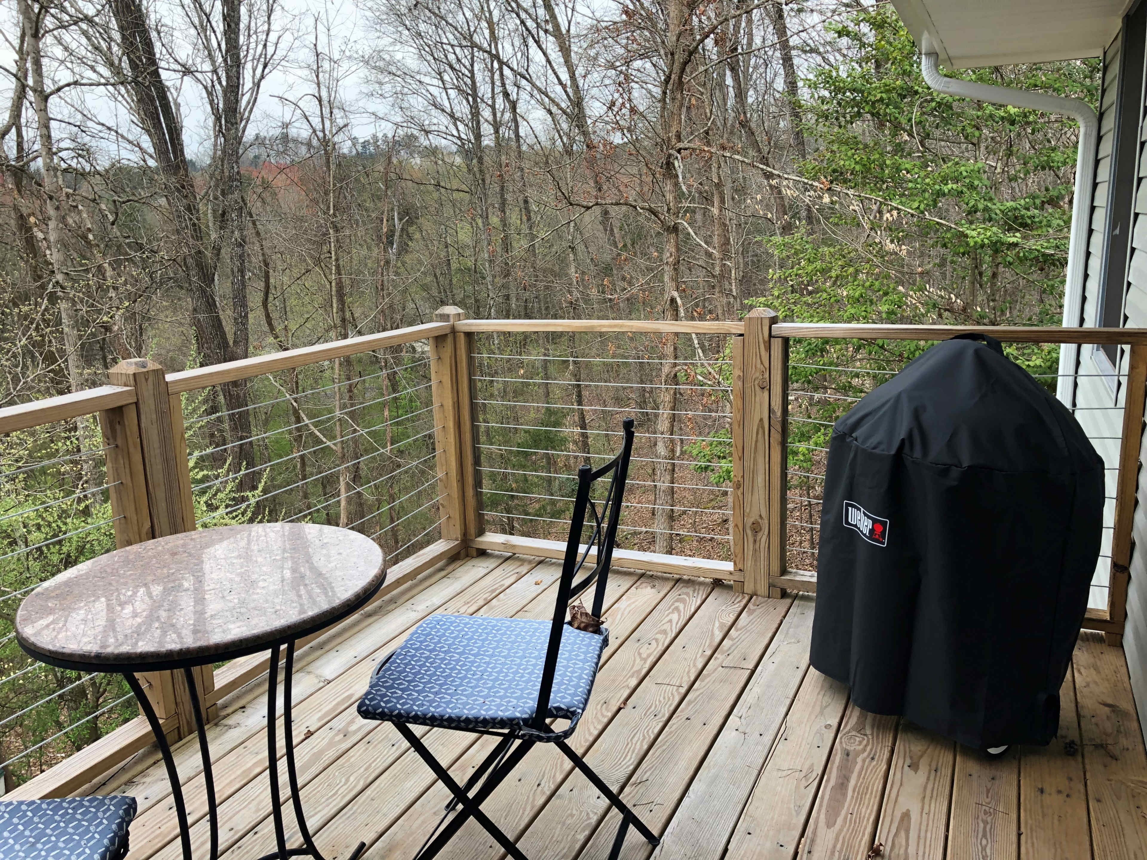 A small wooden balcony with a round table and two chairs, overlooking a wooded area.