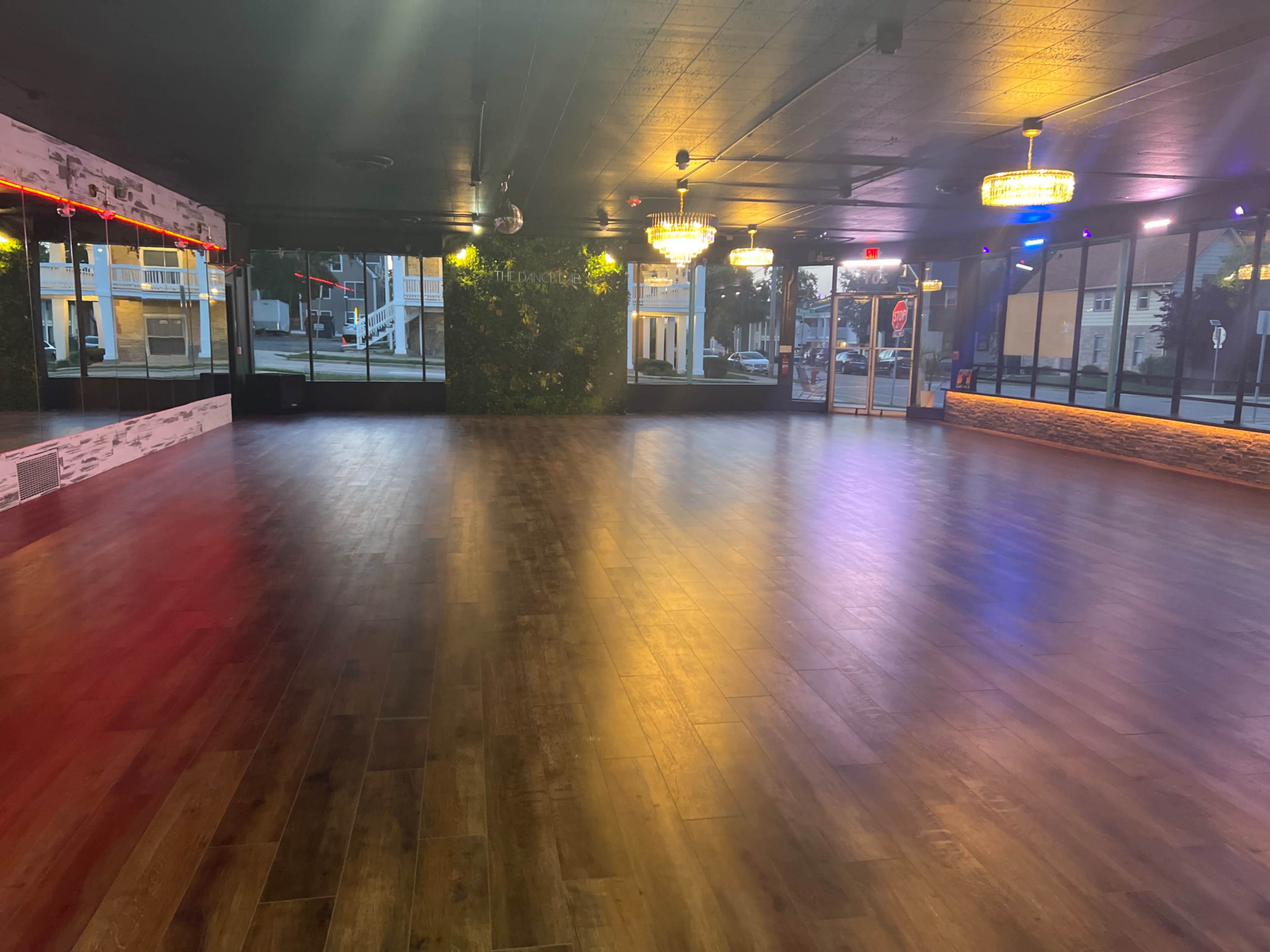 A spacious dance floor with wooden flooring, illuminated by colorful lights and featuring large windows along one side.