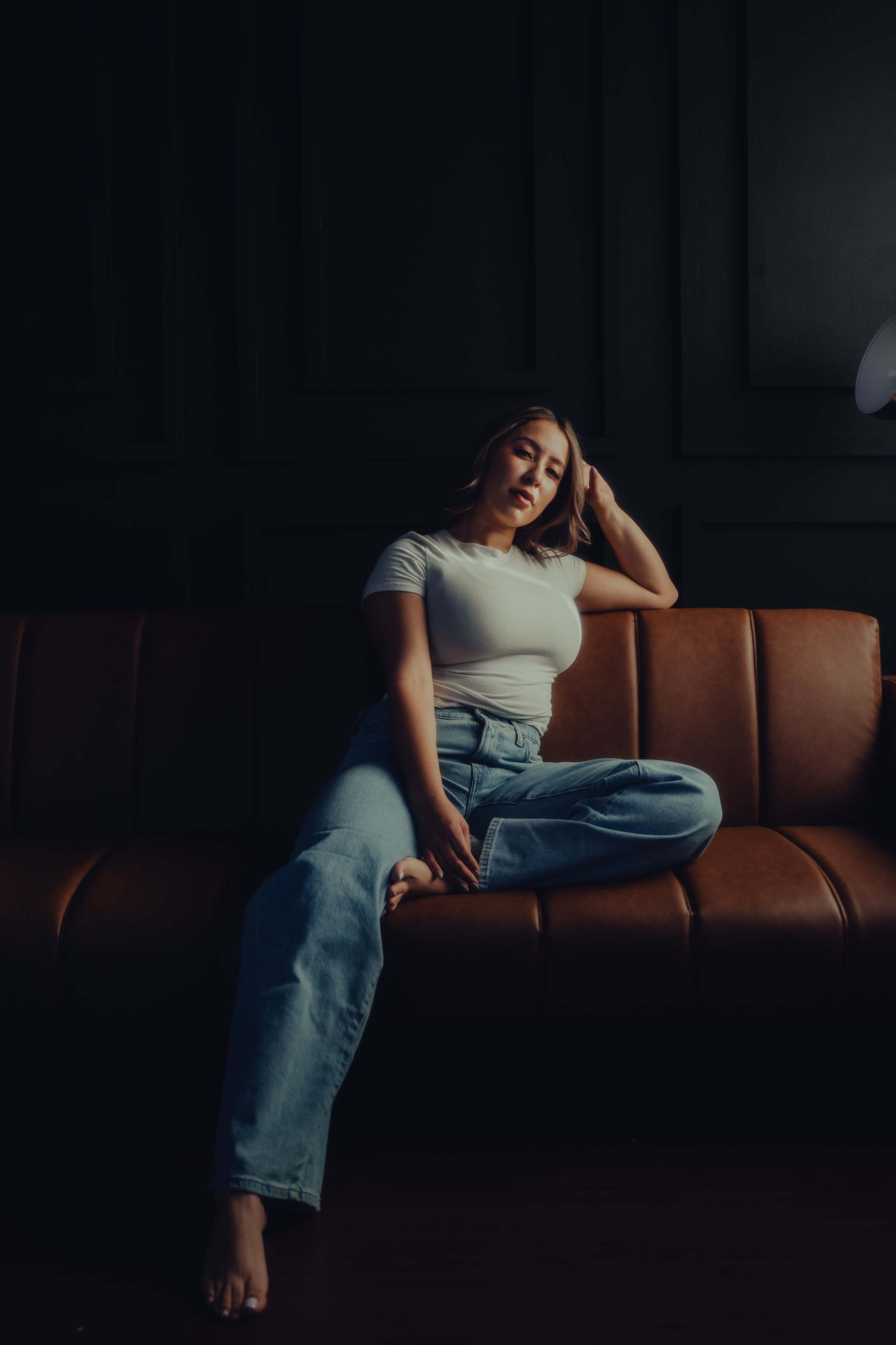 A woman sits on a brown leather sofa in a dimly lit room, wearing a white shirt and blue jeans.