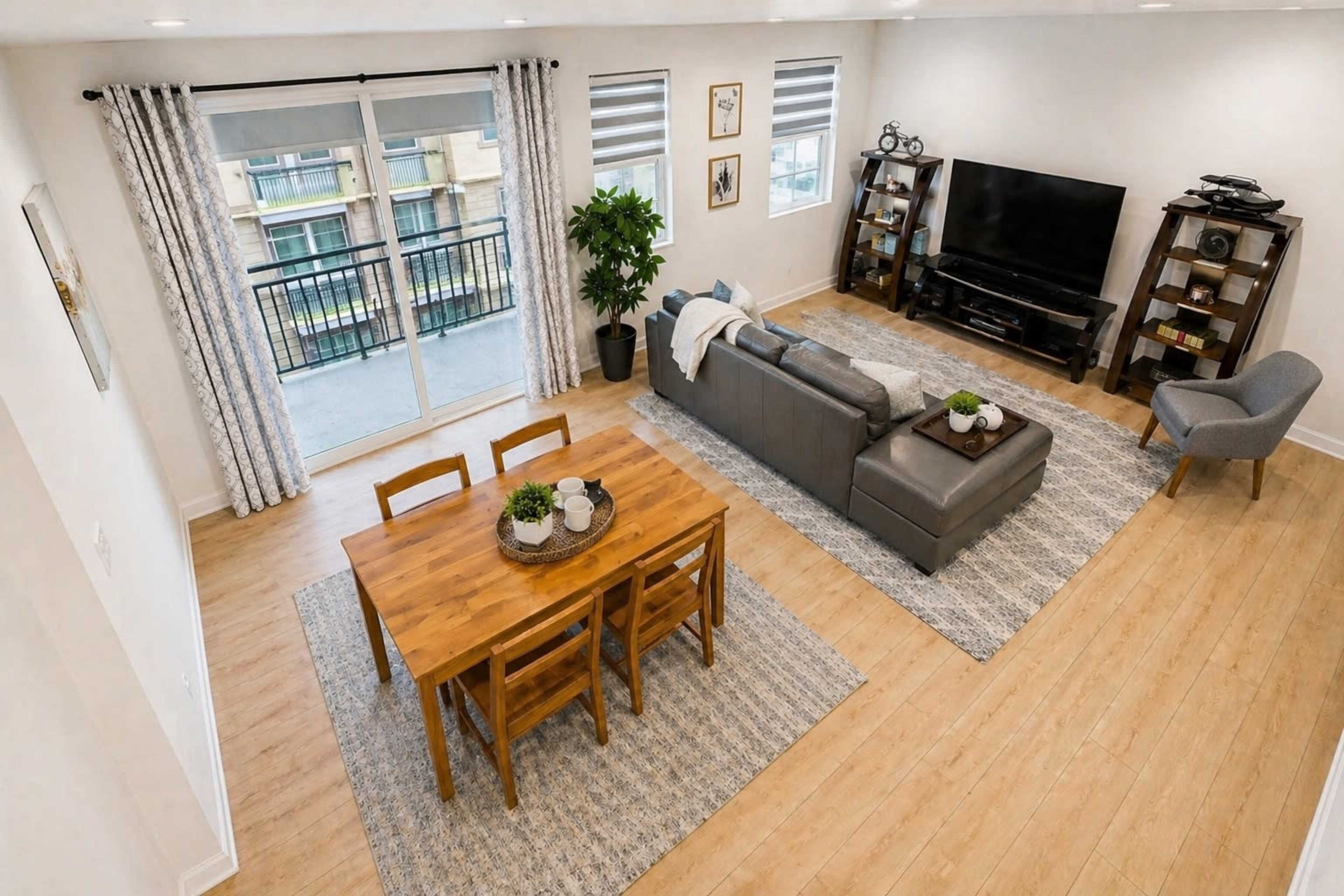 The image shows a modern living space featuring a gray sectional sofa, a wooden dining table with chairs, and a television setup against a wall, with large windows providing natural light.