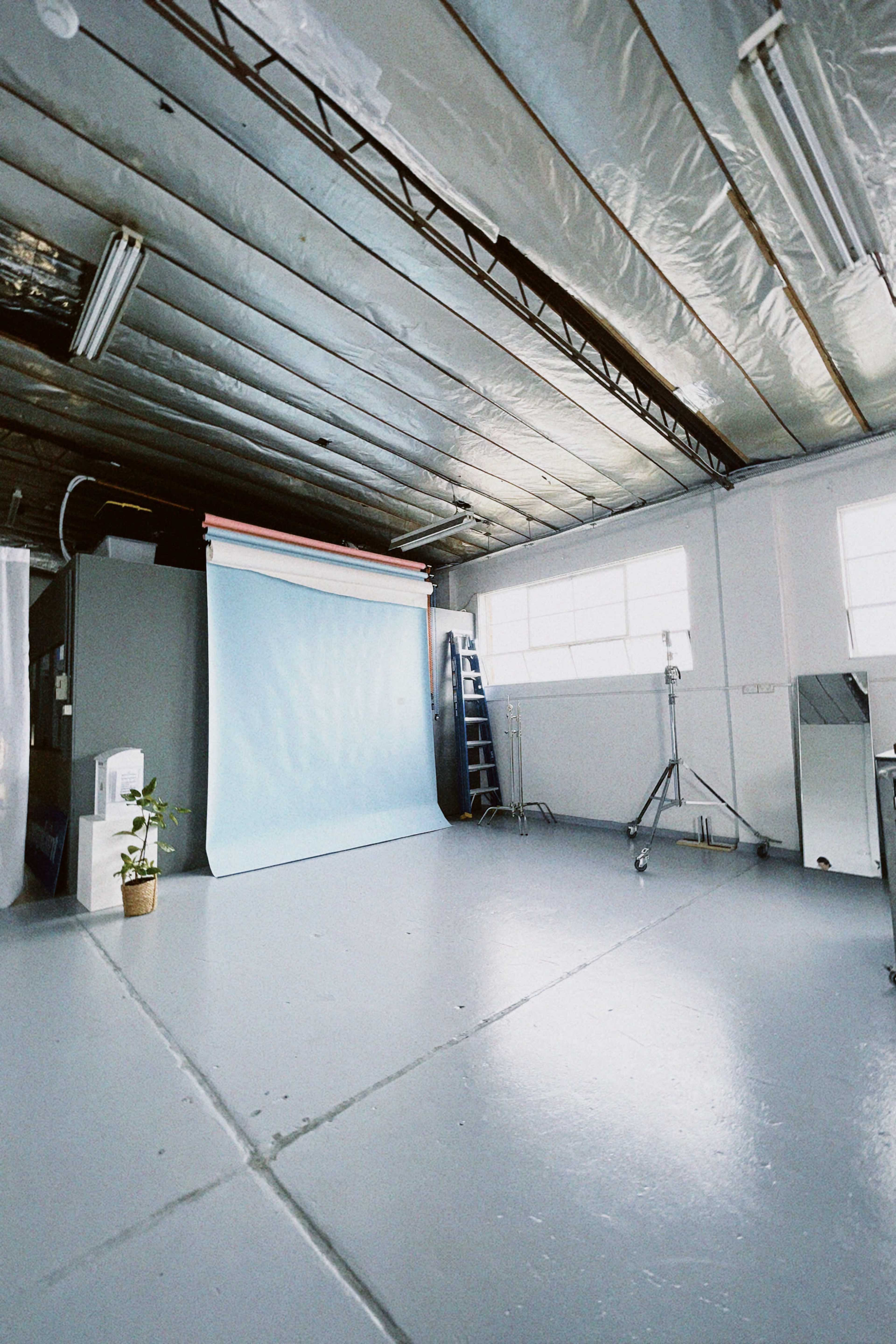 A spacious photography studio with a light blue backdrop, a ladder, lighting equipment, and a potted plant.