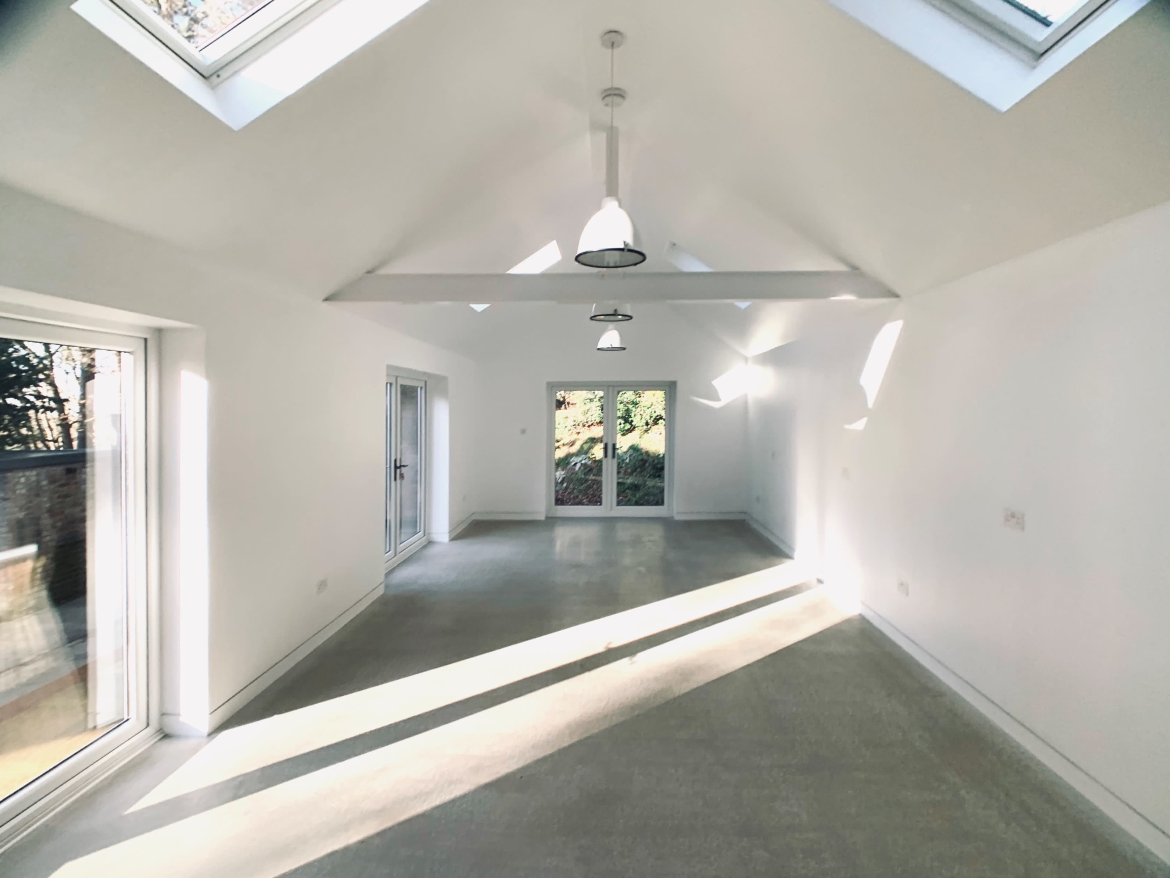The image shows a spacious, brightly lit room with large windows, a vaulted ceiling, and concrete flooring.