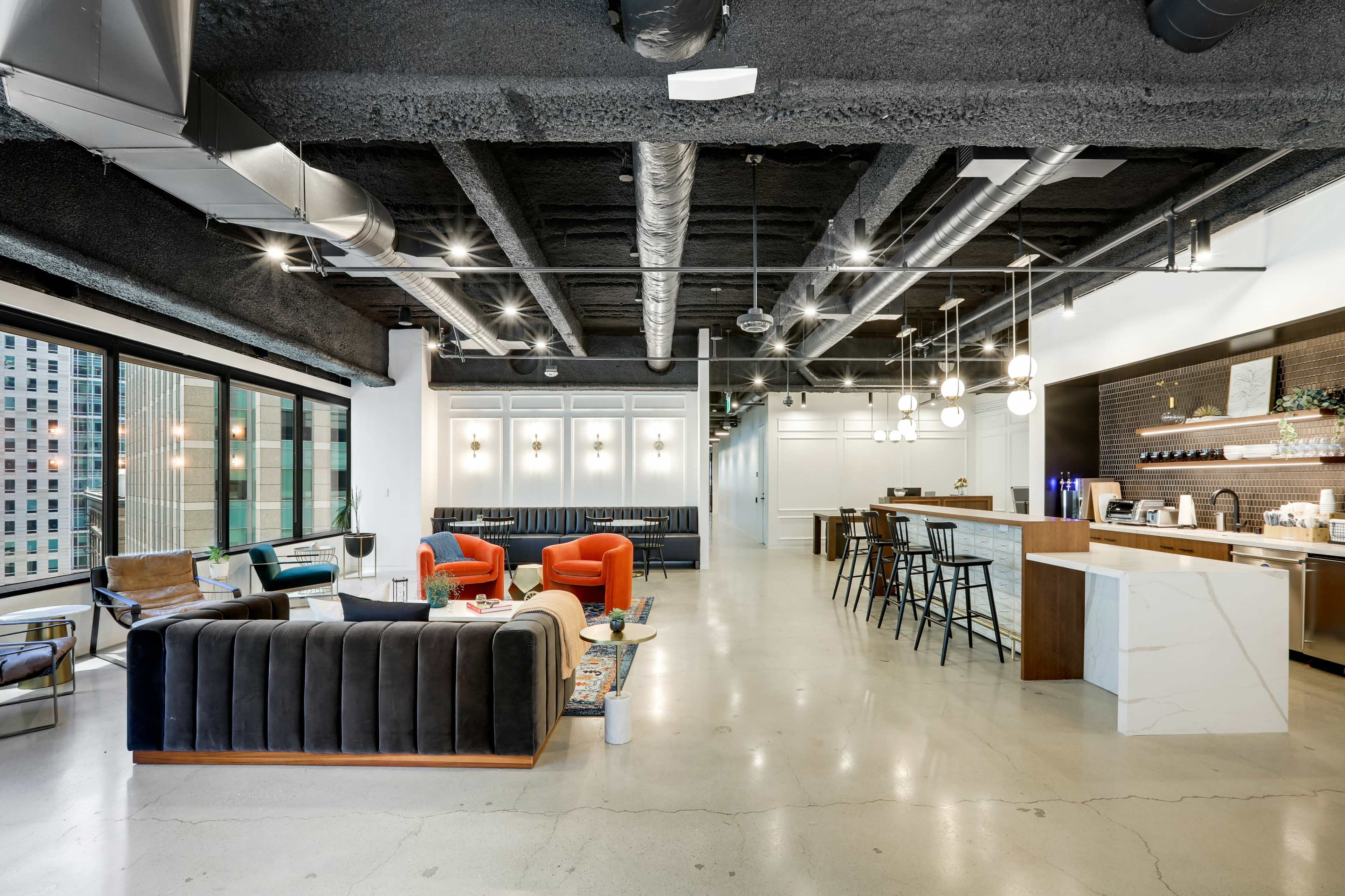 A modern office lounge area with a kitchen, featuring various seating arrangements and large windows.