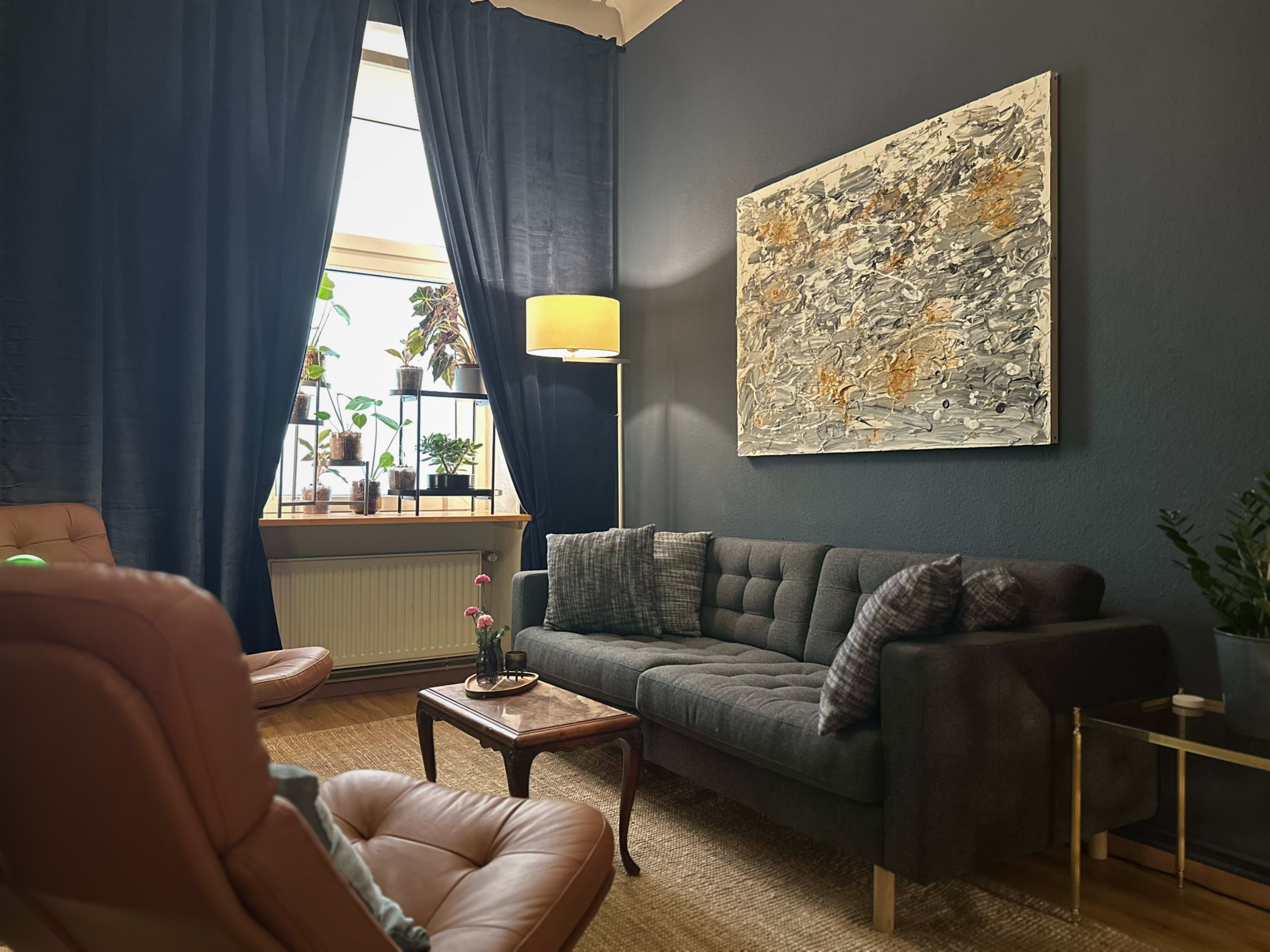 The image shows a cozy living room featuring a dark blue wall, a gray sofa, a wooden table, and large windows with plants.