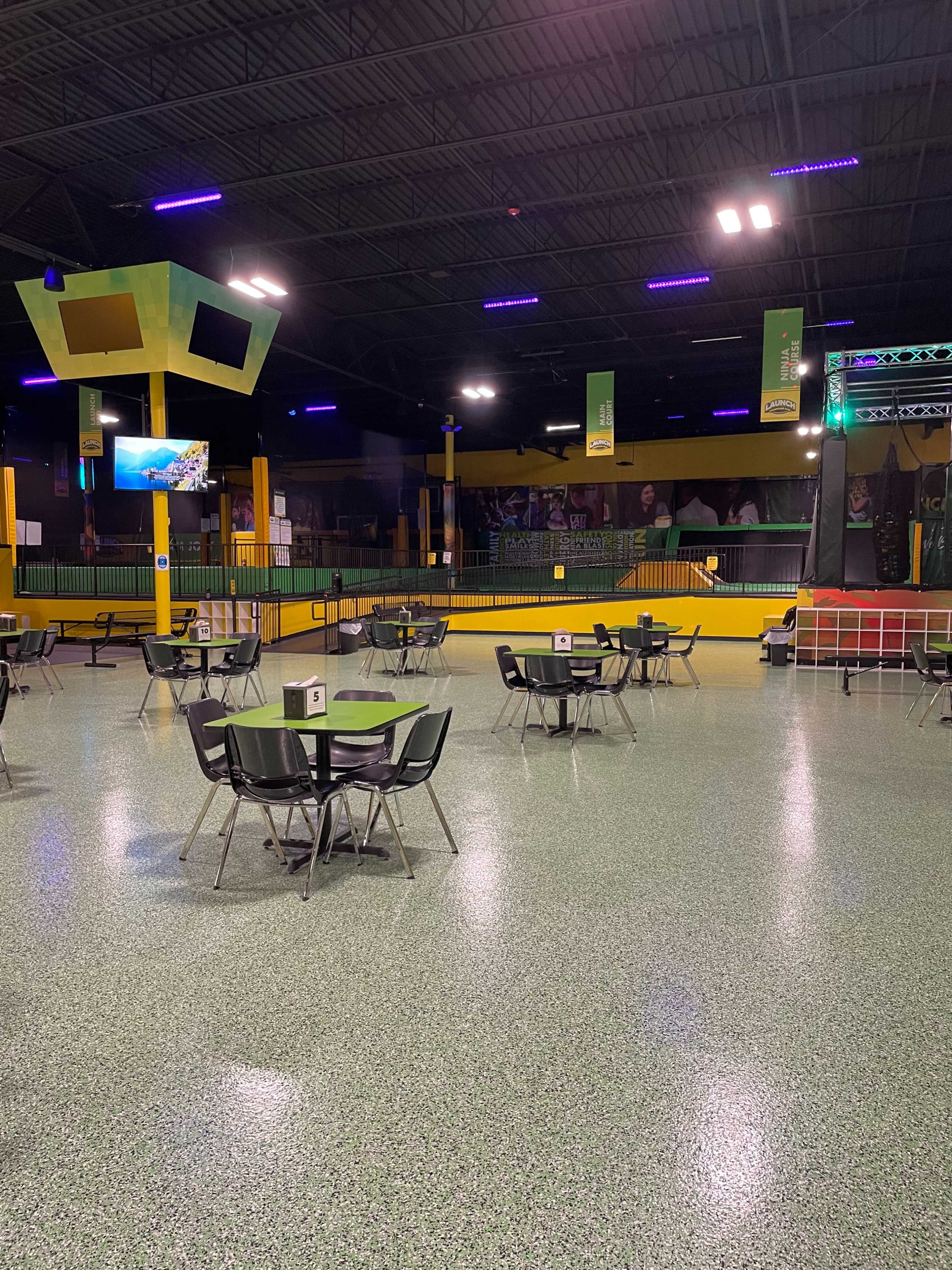 The image shows an empty indoor entertainment space with green and yellow walls, tables, and a large screen.