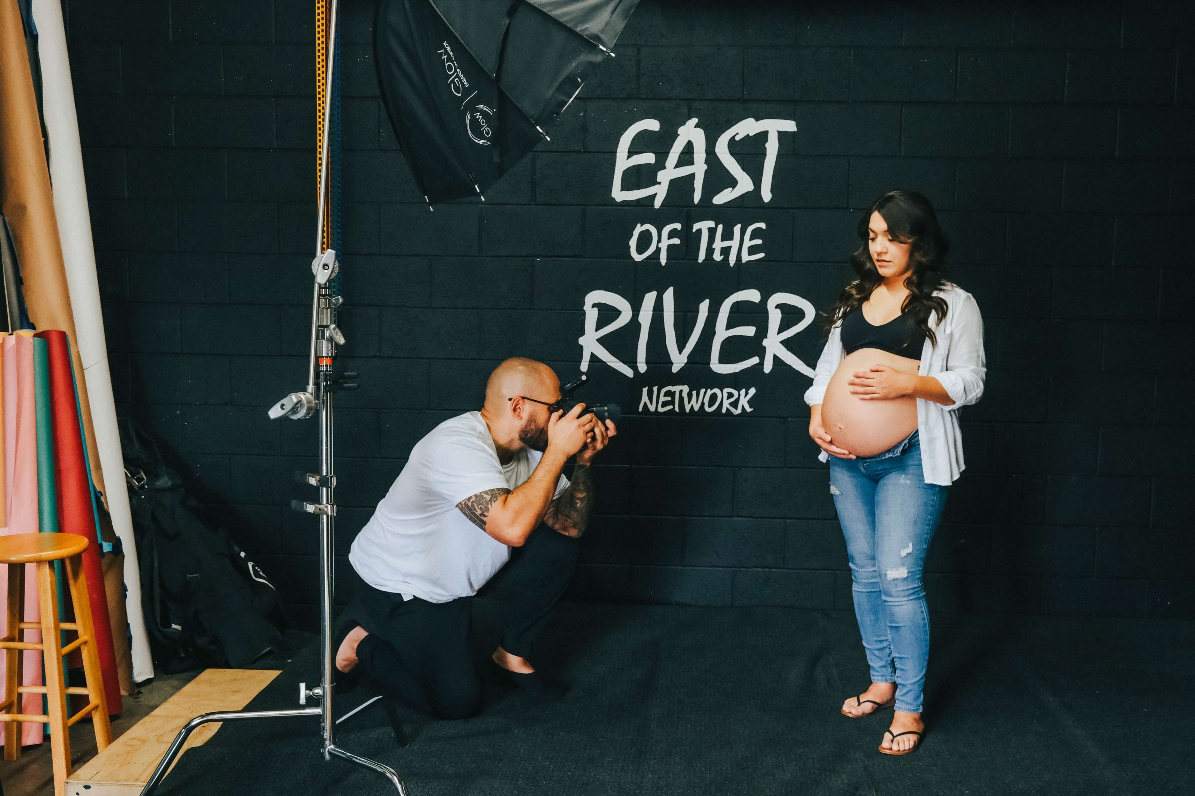 A photographer is capturing a portrait of a pregnant woman in a studio with a black wall featuring the words "EAST OF THE RIVER NETWORK."