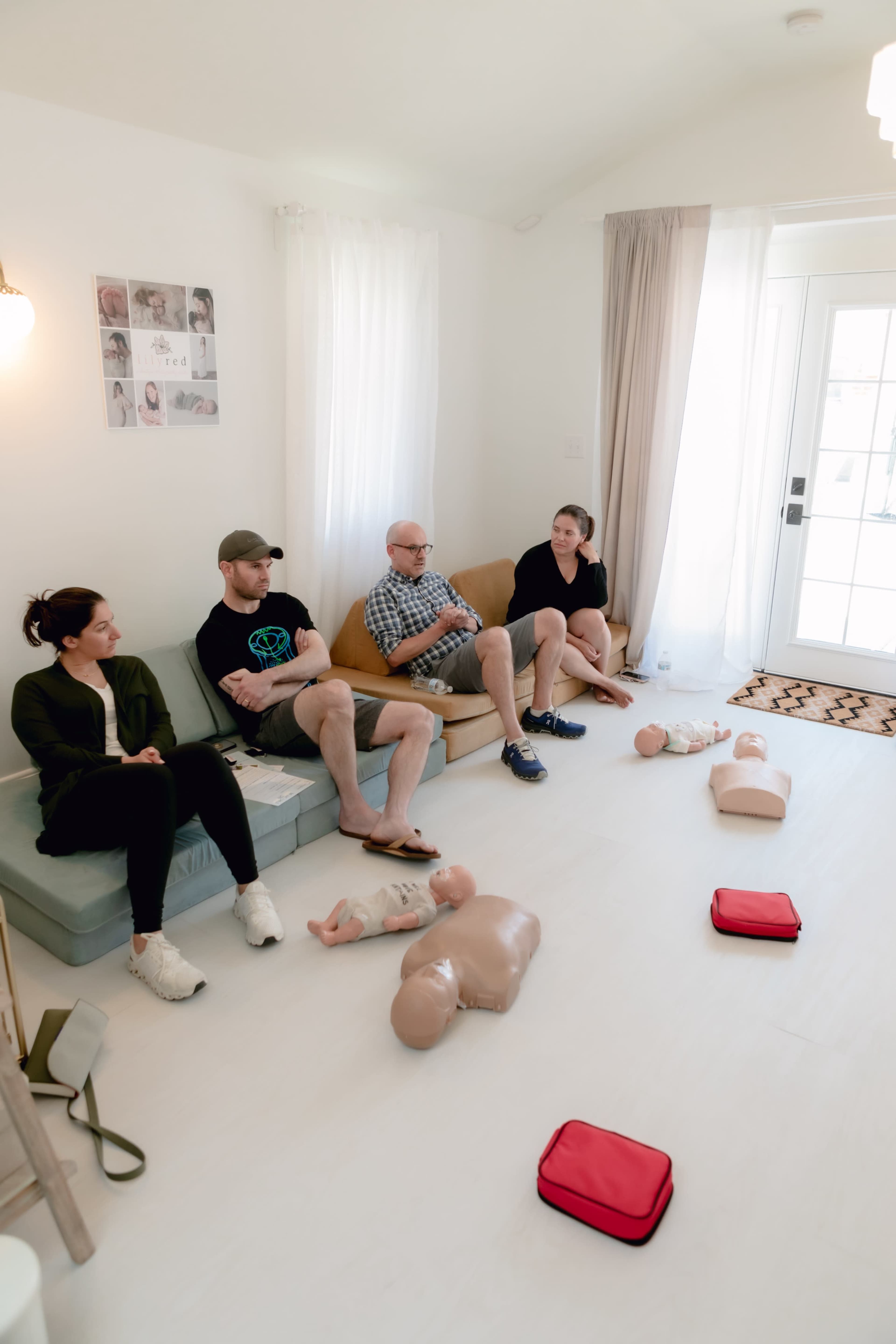 A group of four people sits on a couch in a bright room with CPR mannequins and red first aid kits placed on the floor.