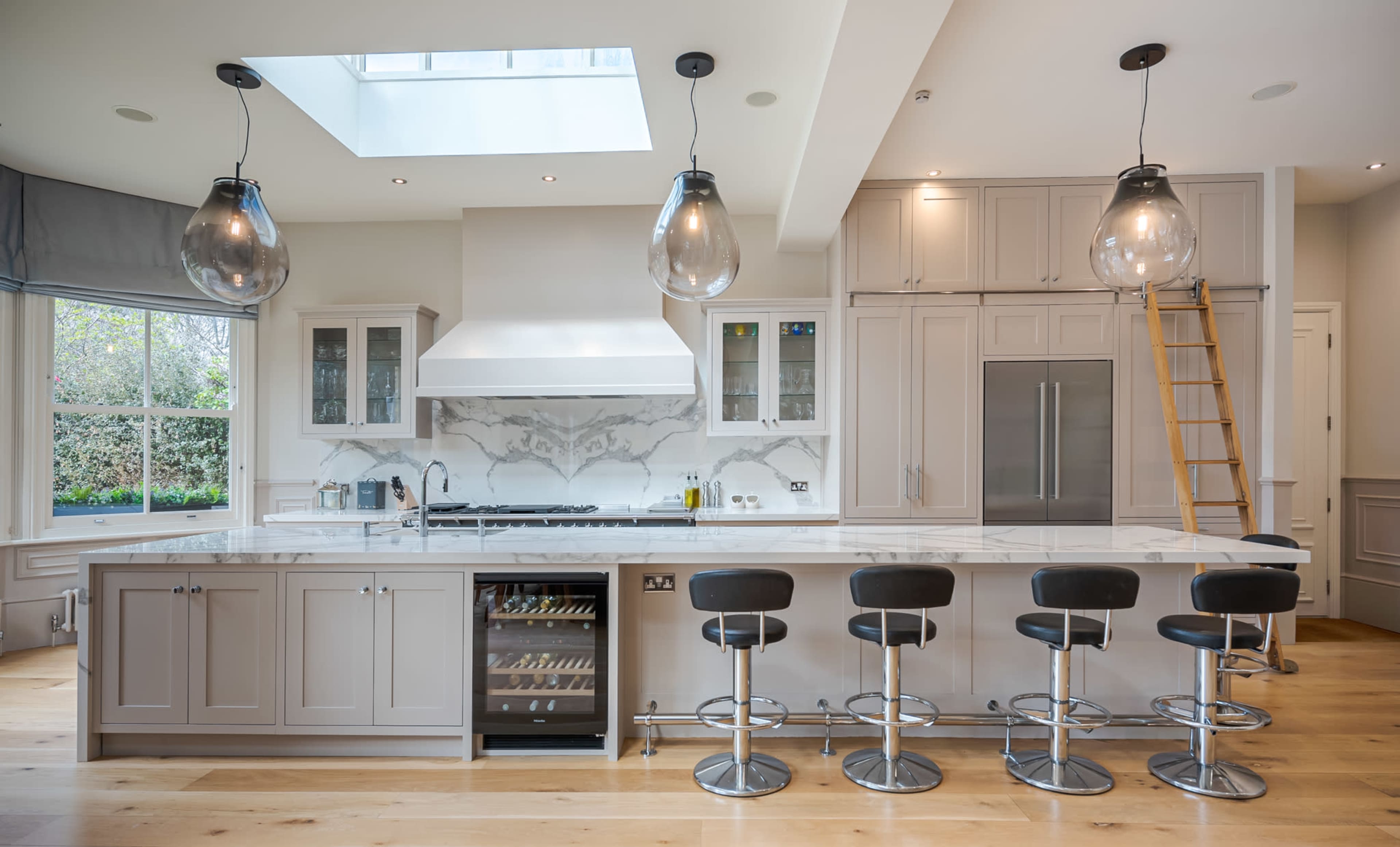 A modern kitchen features a central island with bar stools, a skylight above, and sleek cabinetry alongside a stainless steel refrigerator.
