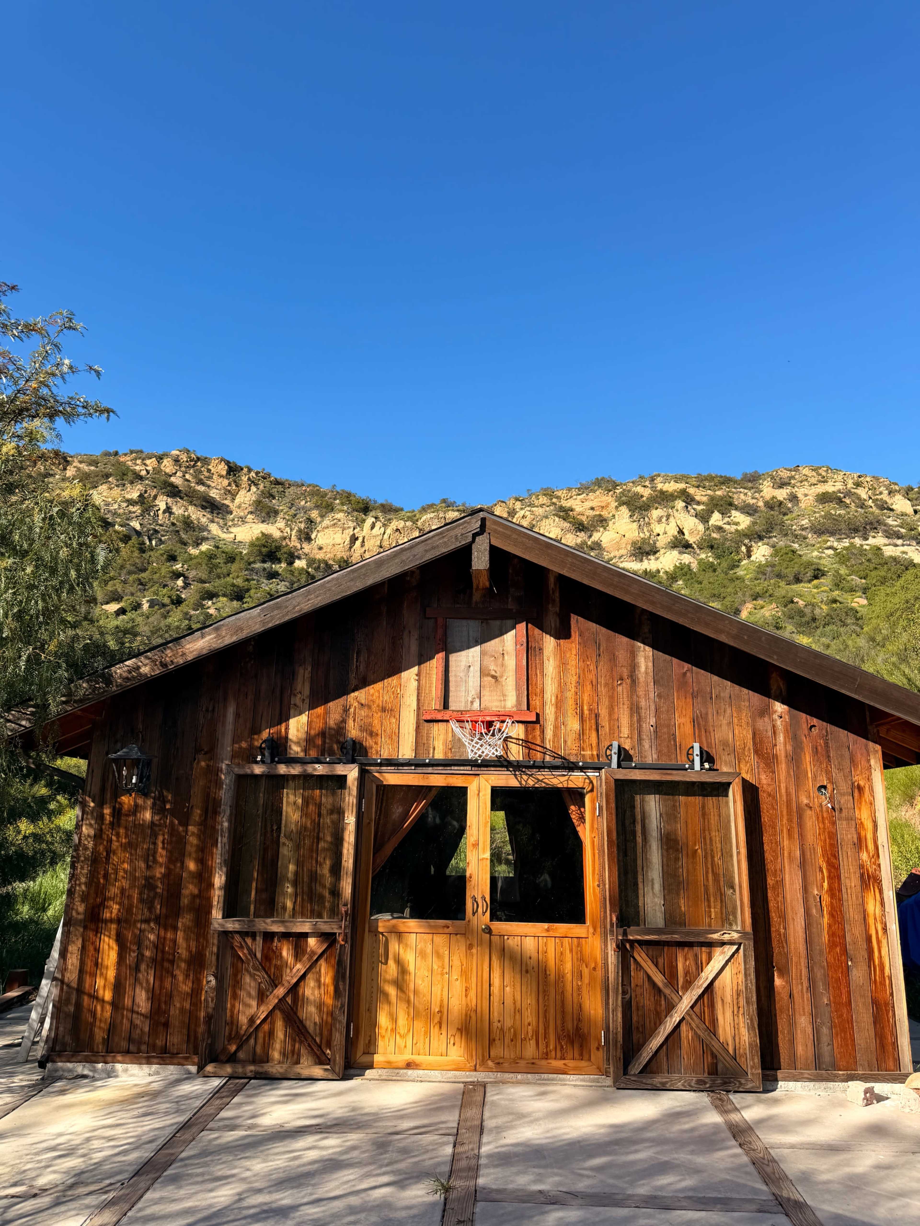 A wooden barn-like structure with a basketball hoop is situated in front of a mountainous landscape under a clear blue sky.