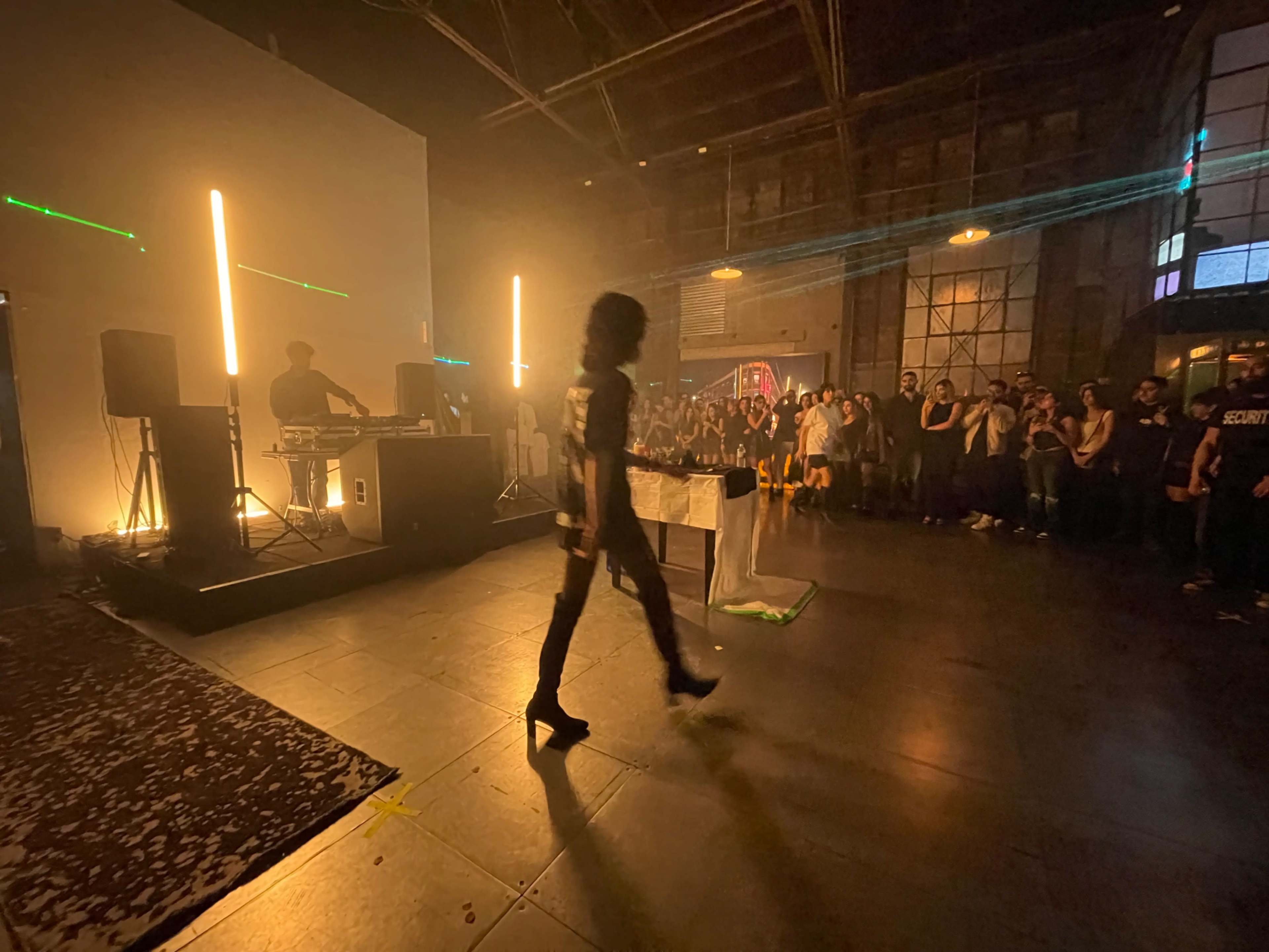 A performer walks onstage in front of an audience while a DJ plays music in a dimly lit venue.