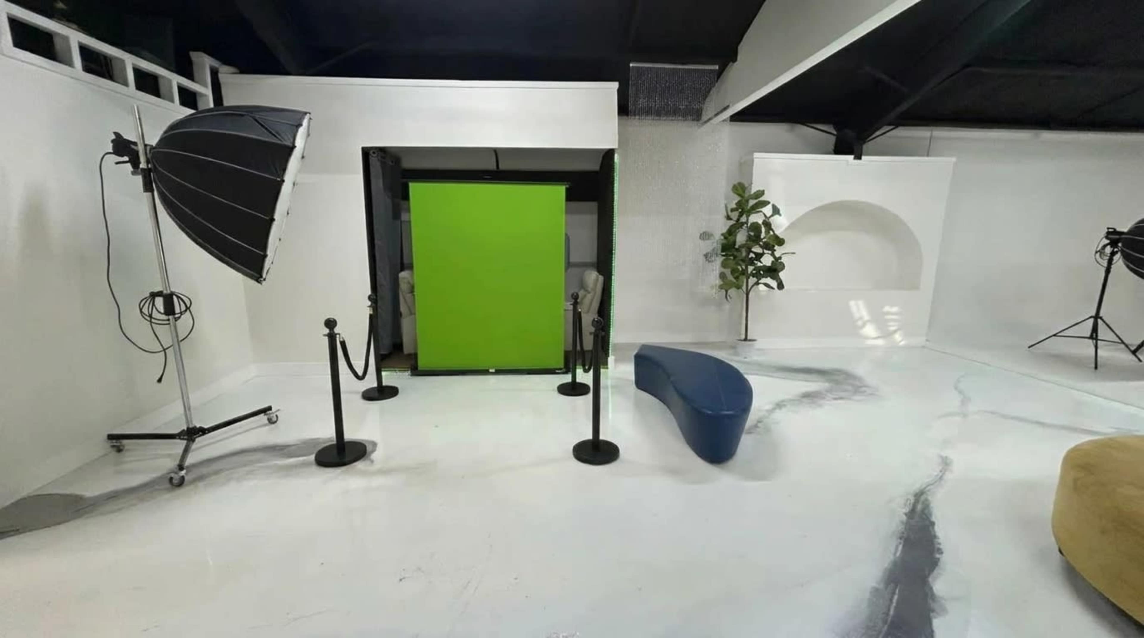 The image shows a photography studio with a green screen in the center, surrounded by lighting equipment, a decorative plant, and various props.