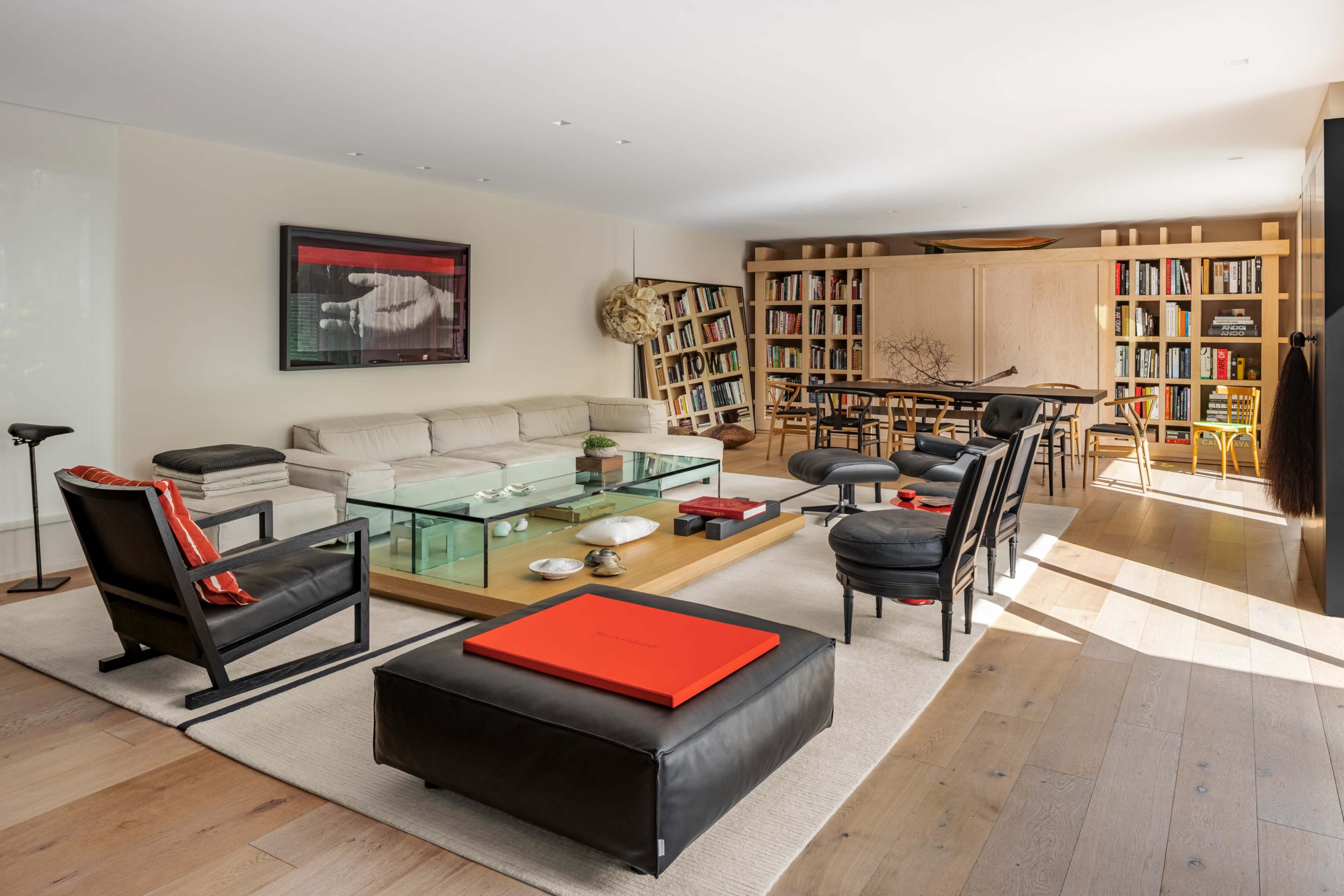 The image shows a modern living room featuring a large beige sofa, a wooden coffee table, black chairs, and a bookshelf filled with books along one wall.